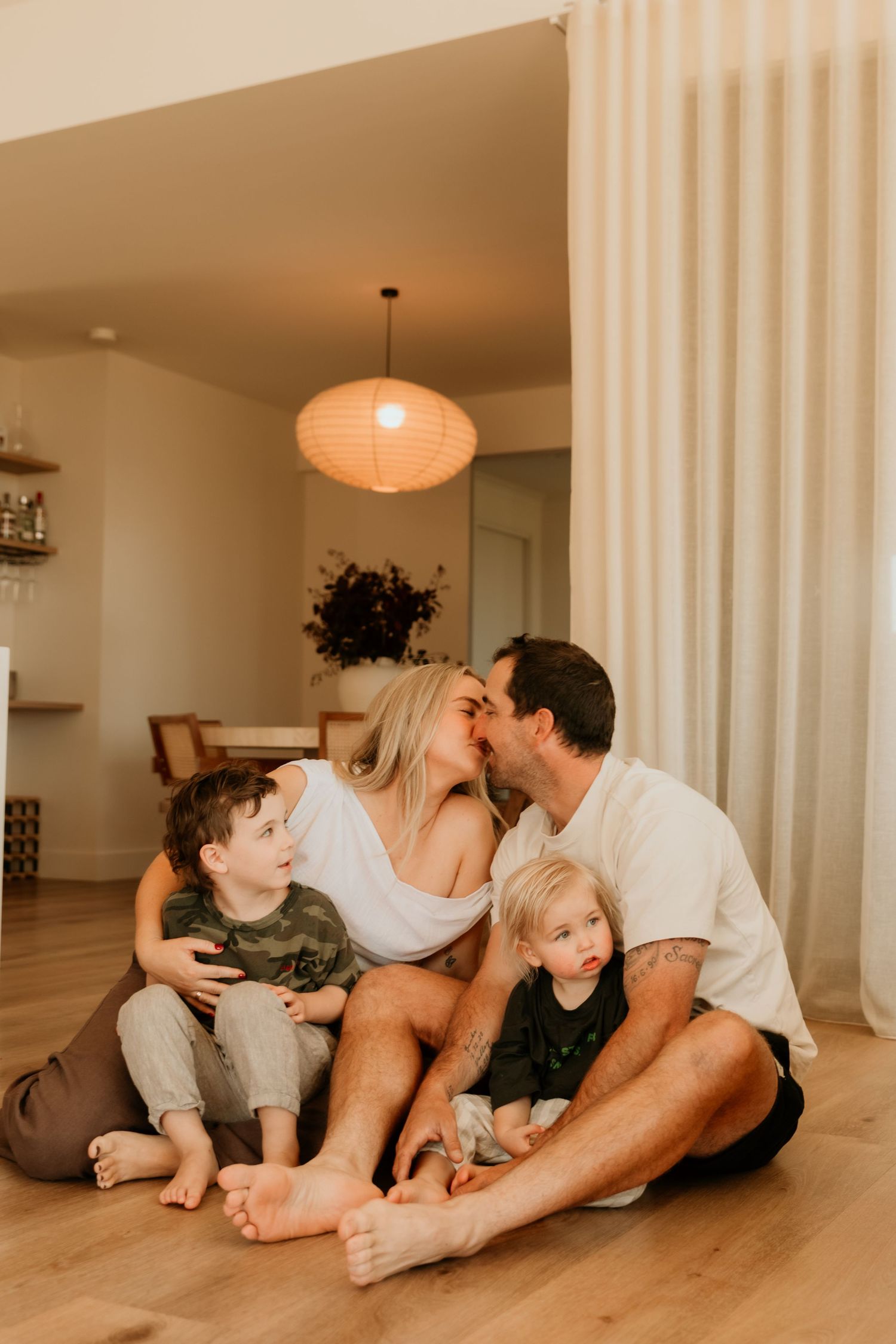 Parents kissing while seated on floor with two young children in warmly lit modern living room.