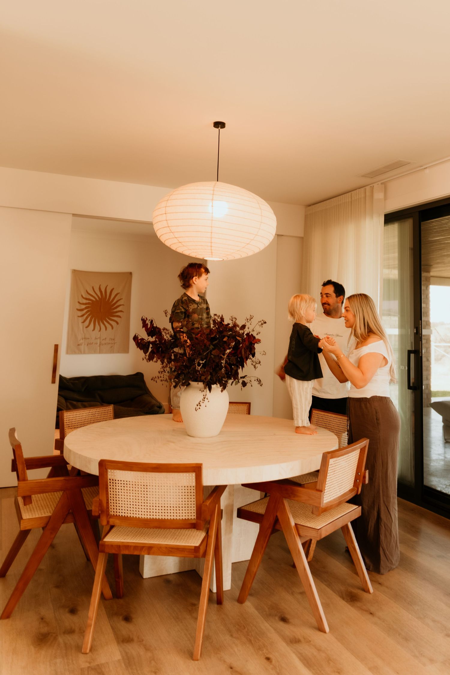 Family gathered around a round dining table with rattan chairs beneath a paper lantern pendant light in a modern home.