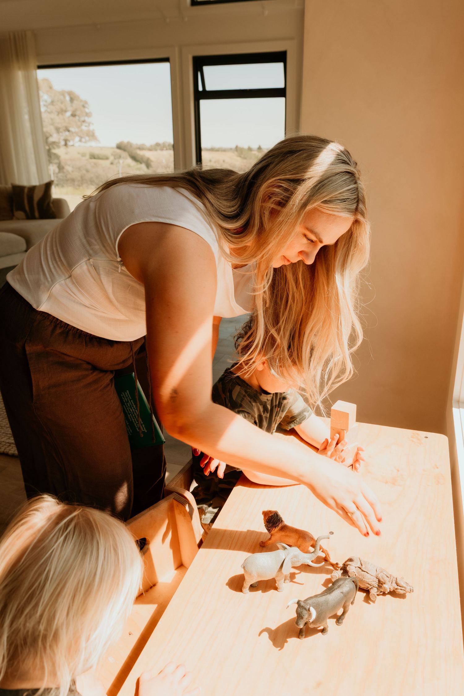 Blonde mother in white top playing with animal figurines on a wooden table with her young child in sunny room.