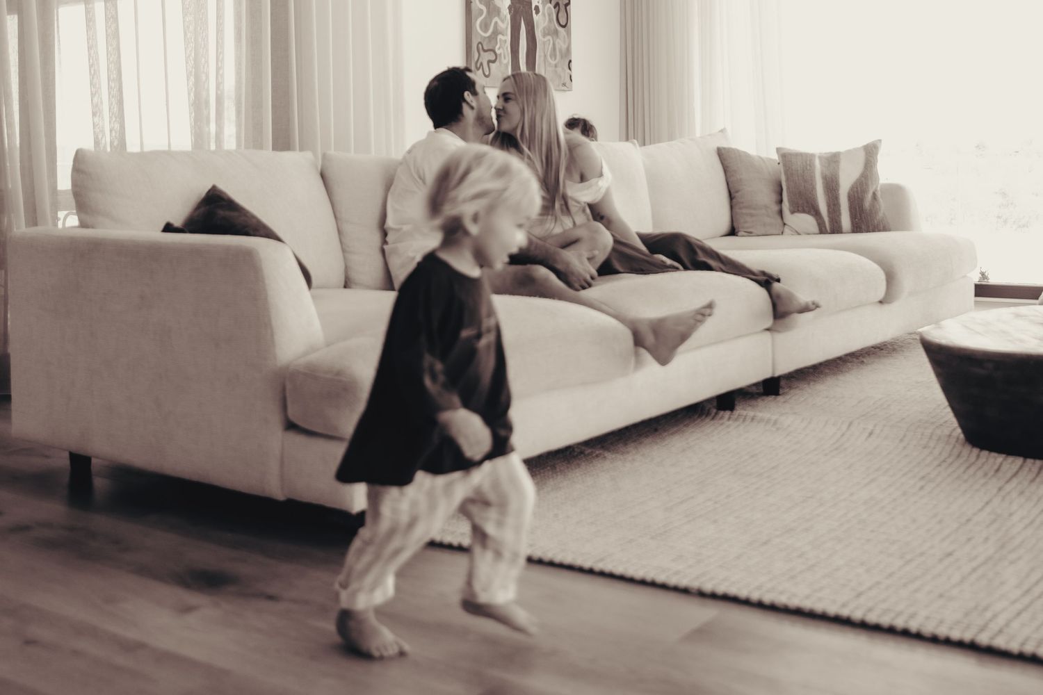 Black and white photo of a family relaxing on a white sofa while a child runs playfully in foreground.