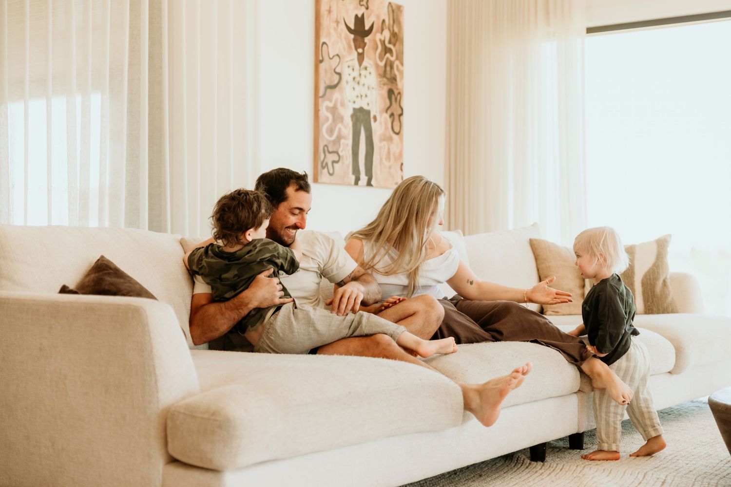 Happy family of four laughing and cuddling together on a white couch in a bright modern living room.