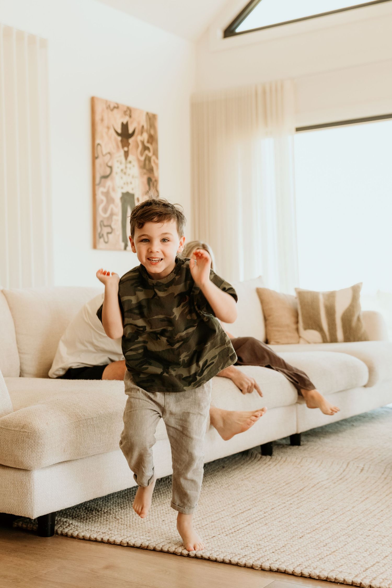 Energetic young boy in camo shirt jumping barefoot on a rug in a bright modern living room.