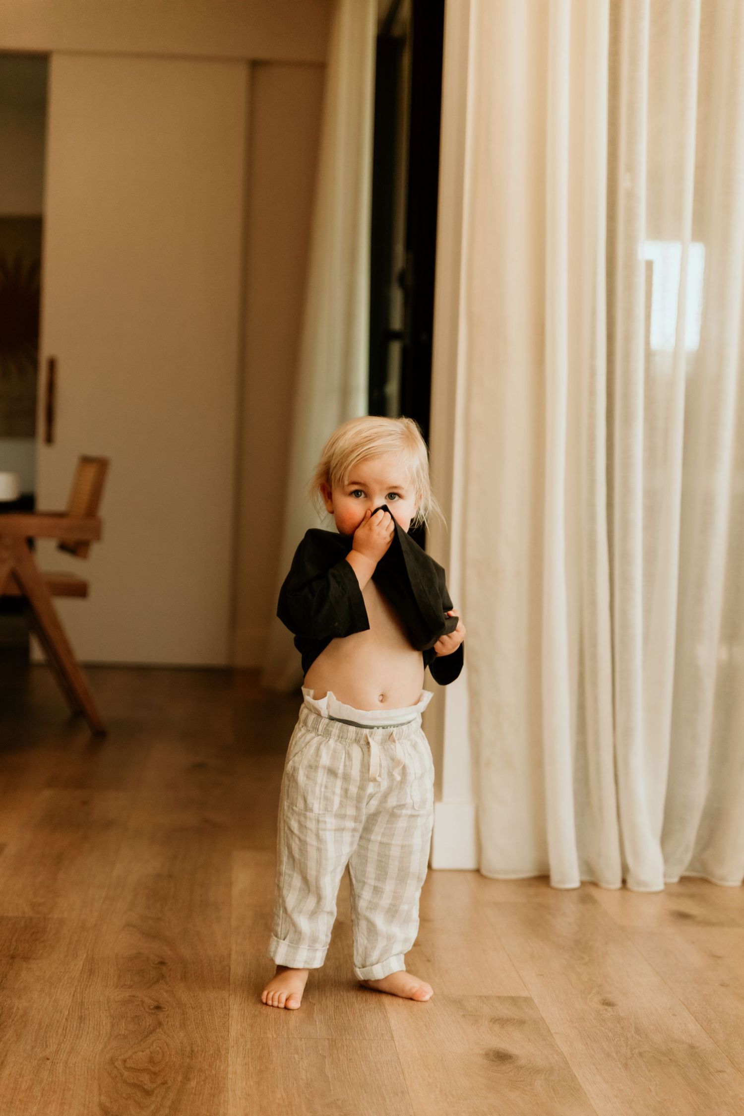 Adorable blonde toddler standing barefoot on hardwood floor lifting shirt in a bright modern home.