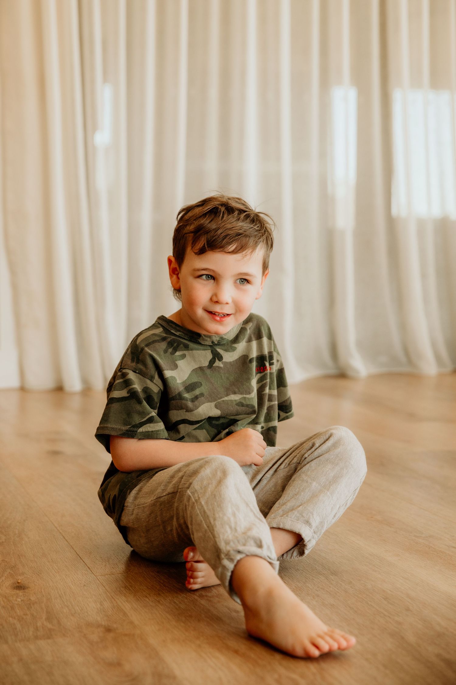 Smiling young boy in camo shirt sitting cross-legged barefoot on hardwood floor near white curtains.