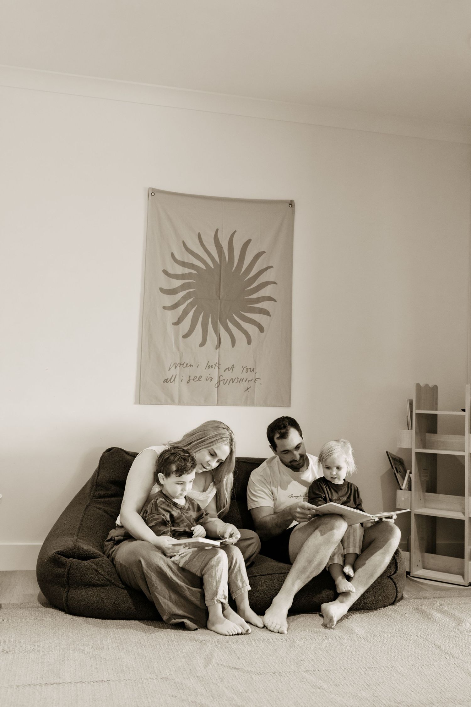 Family of four sitting together on bean bag chairs reading a book beneath a sun wall tapestry in black and white.