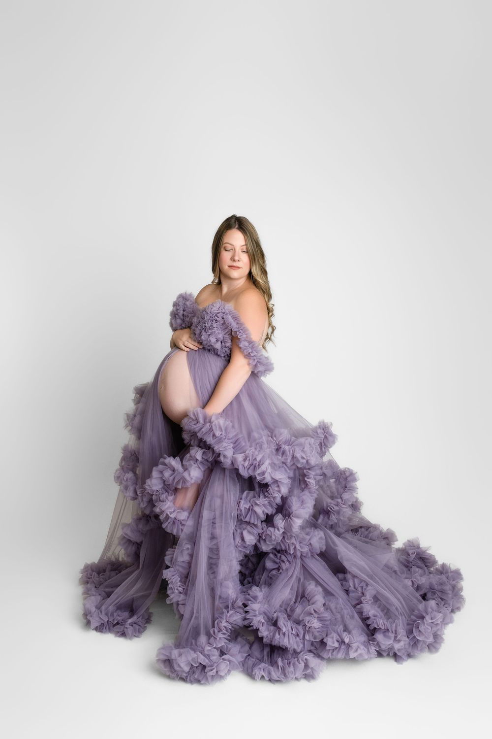 A pregnant woman poses in a flowing lavender tulle gown with ruffled details against a white background.