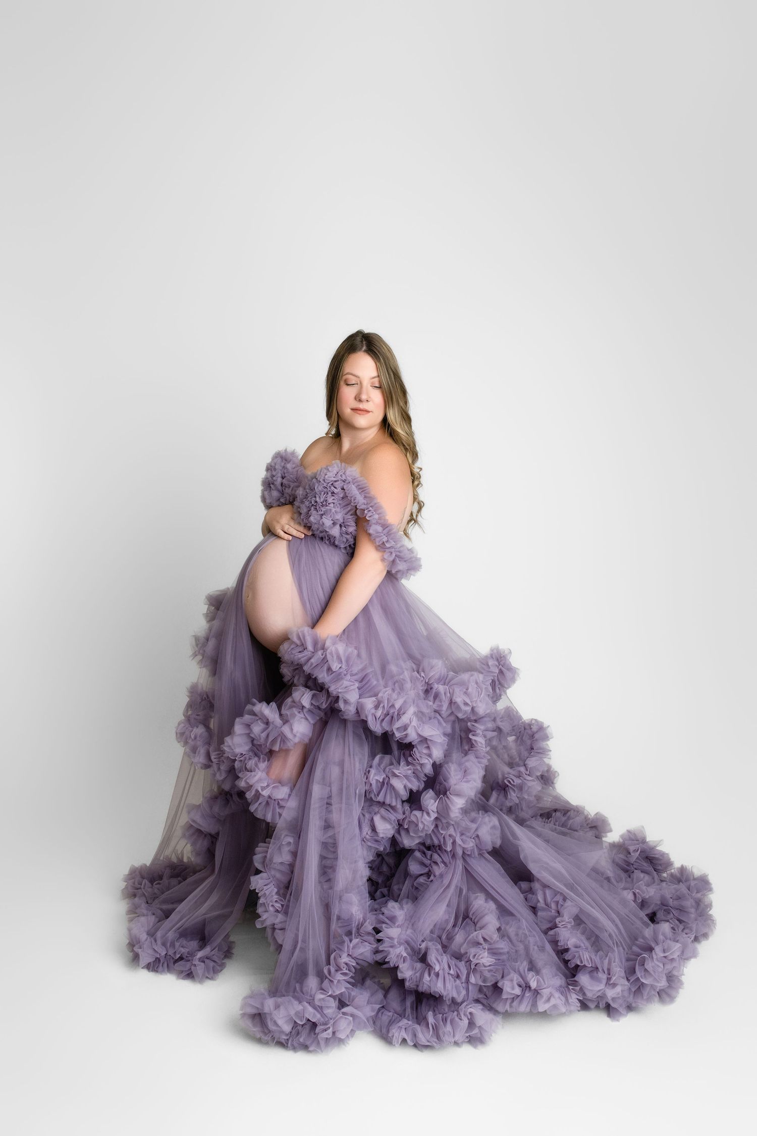 A pregnant woman poses in a flowing lavender tulle gown with ruffled details against a white background.