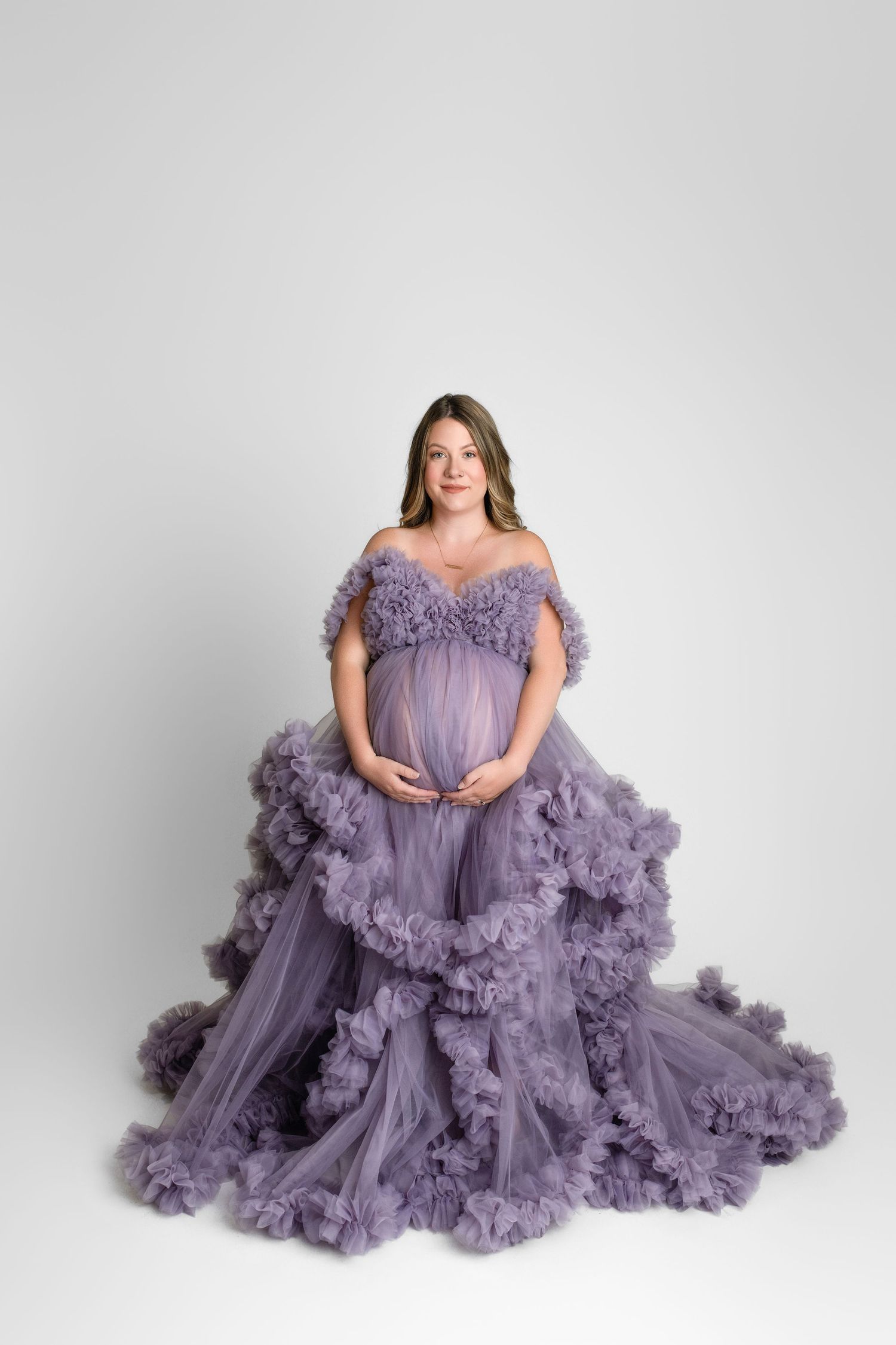 Elegant lavender tulle maternity gown with ruffled layers and off-shoulder neckline photographed against white backdrop.