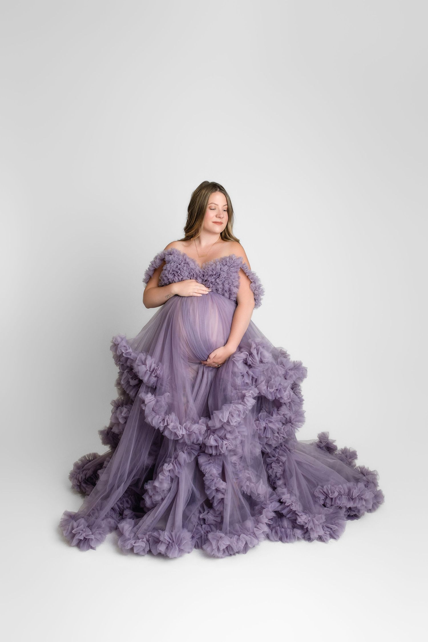 A pregnant woman poses in a flowing lavender tulle gown with ruffled details against a white studio backdrop.