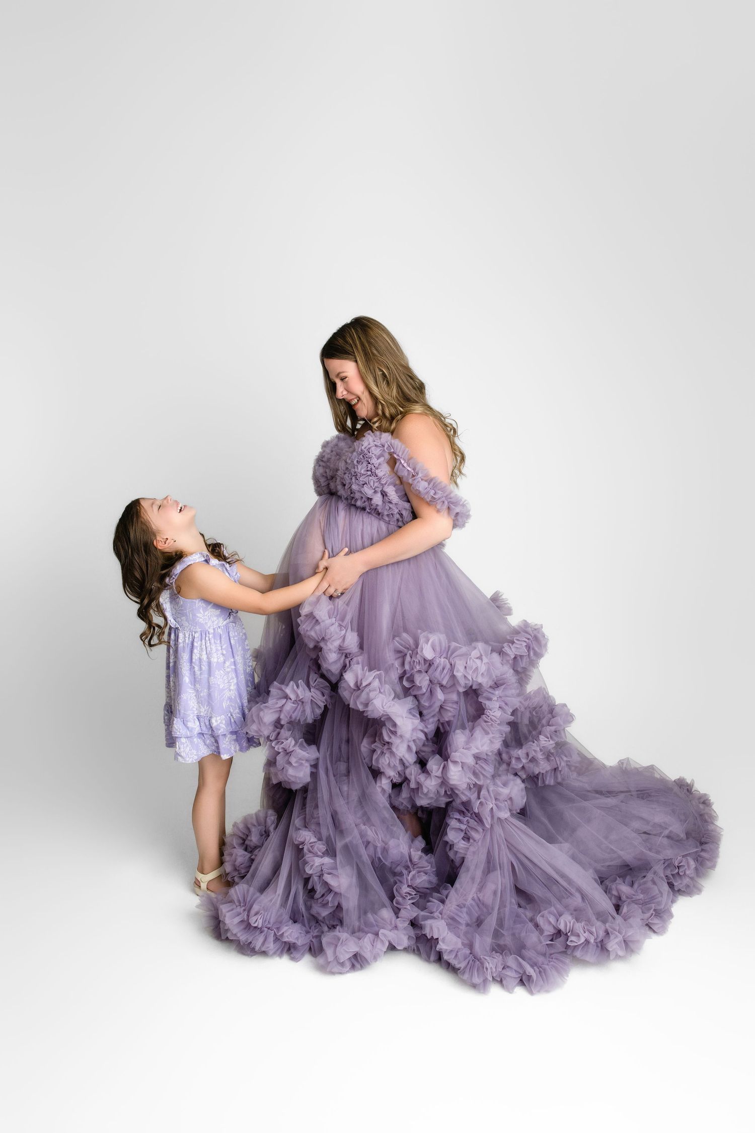 Two people in flowing lavender tulle dresses pose together against a white backdrop in a studio setting.