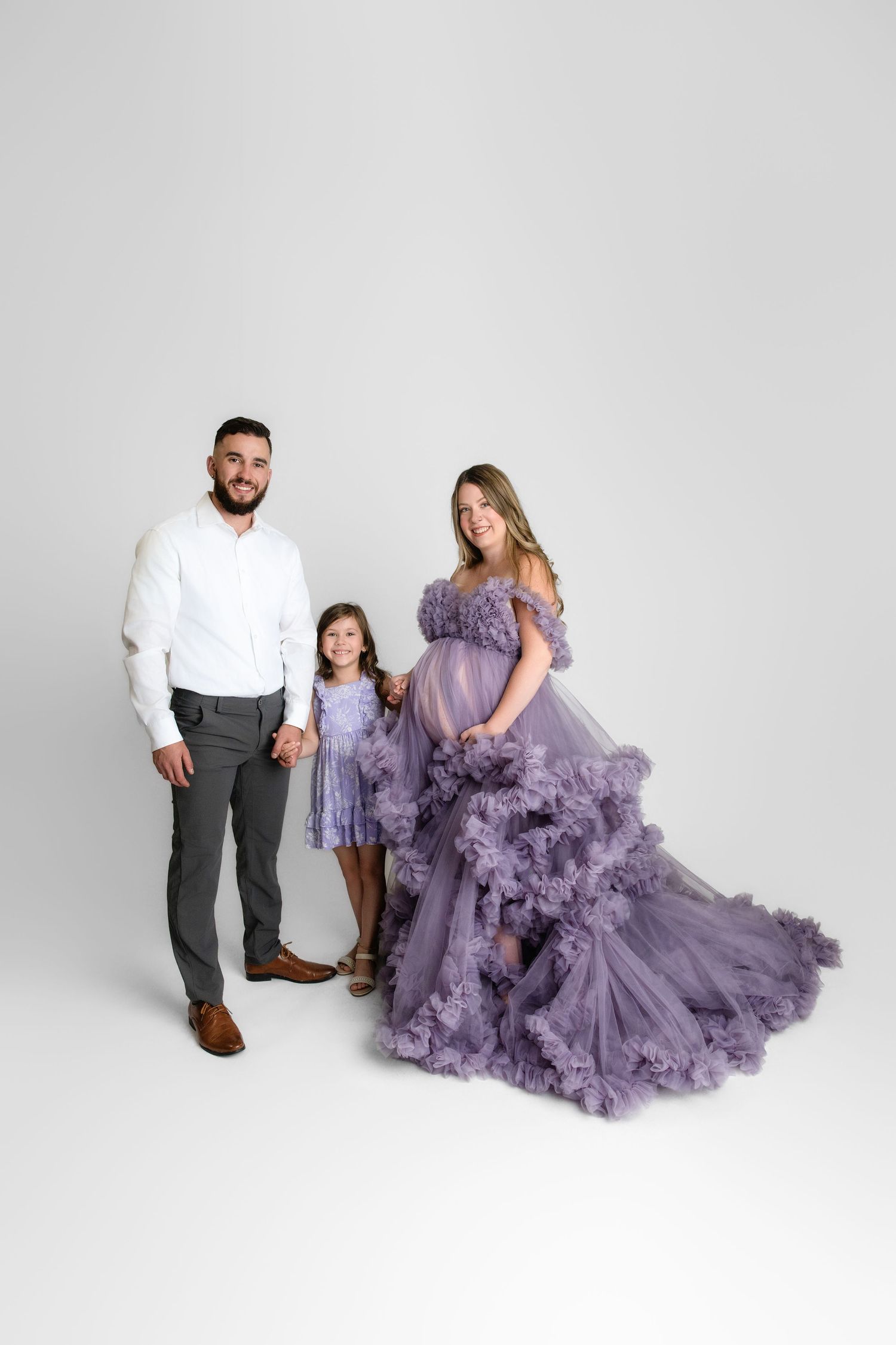 Family portrait in coordinating outfits featuring a dramatic purple tulle gown and white and gray formal wear against white backdrop.