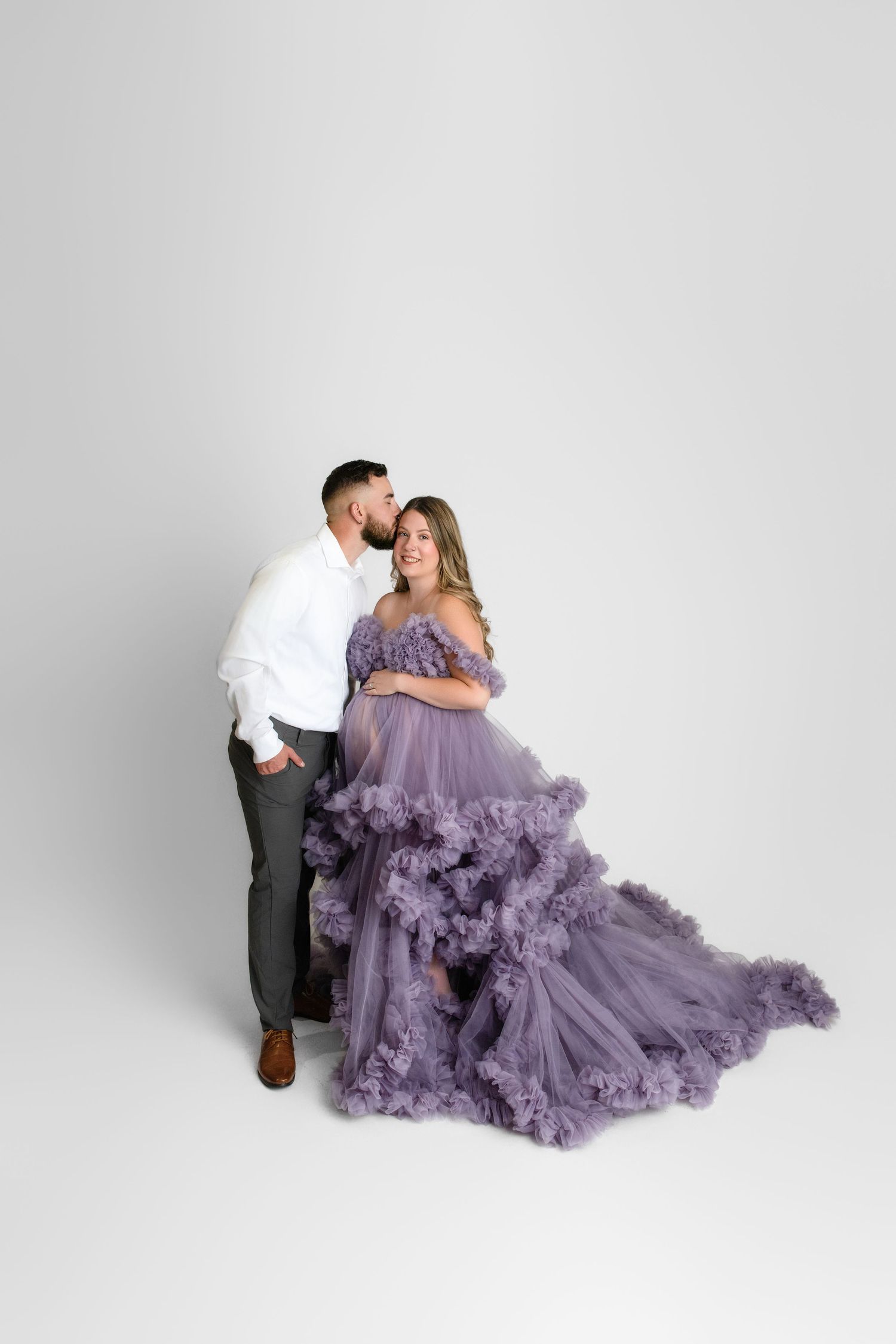 A couple poses together against white backdrop with woman wearing flowing purple tulle maternity gown.