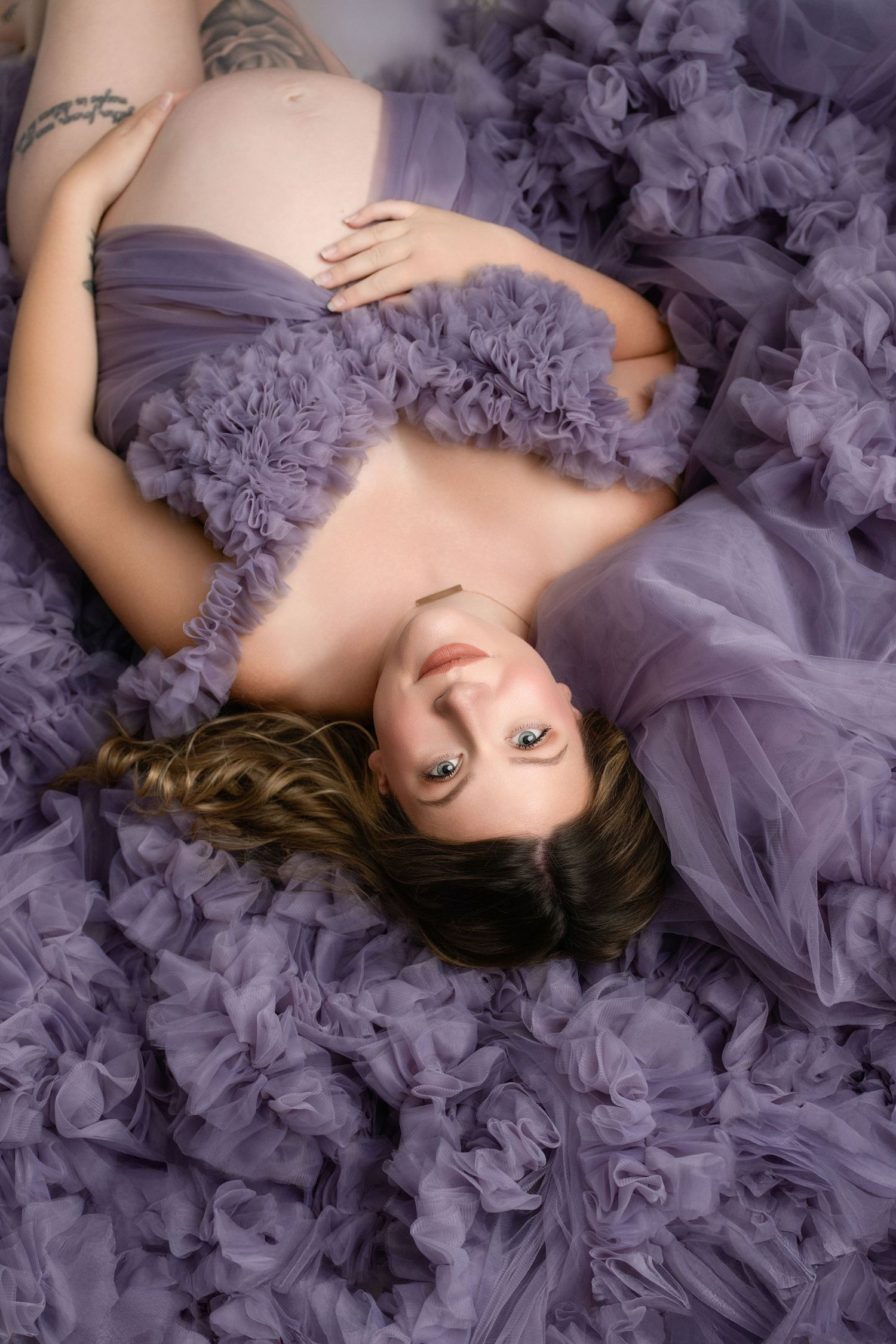 Woman in lavender tulle gown lying on white backdrop during maternity photoshoot.