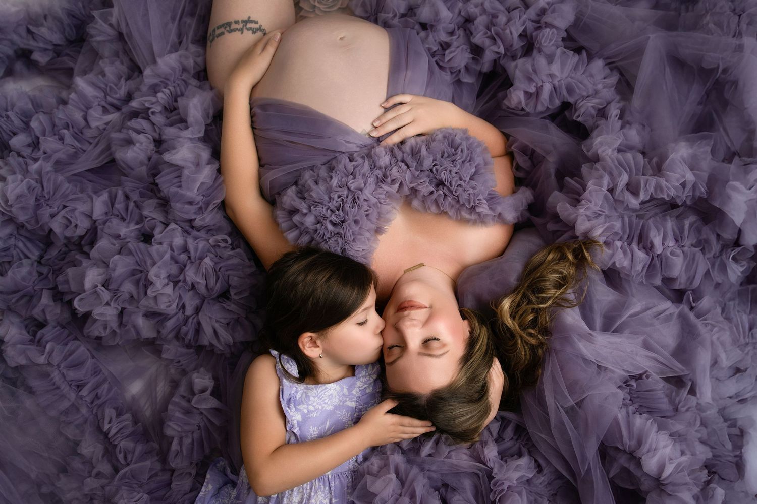Mother and daughter in matching purple tulle dresses lay on floor while sharing tender moment together.