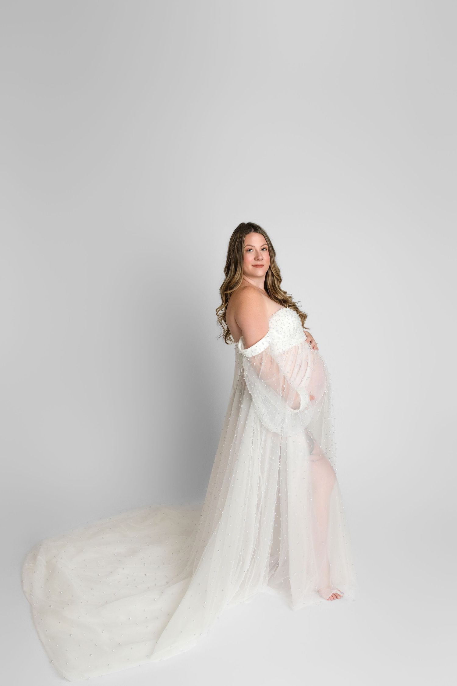 Ethereal maternity photo in flowing white gown with long train against light gray studio backdrop.