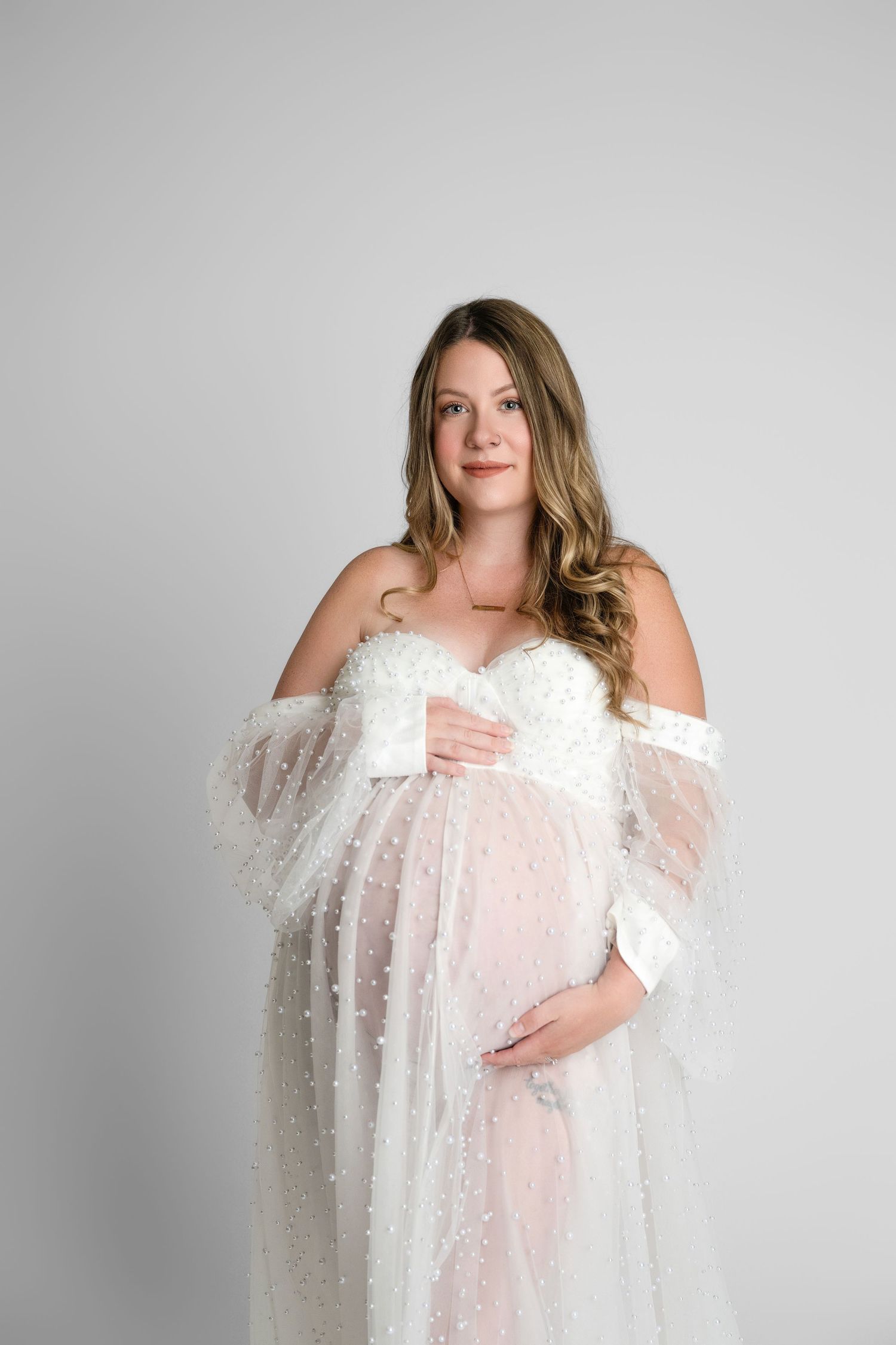 A pregnant woman wearing a white lace maternity gown poses against a light gray backdrop during a studio photoshoot.