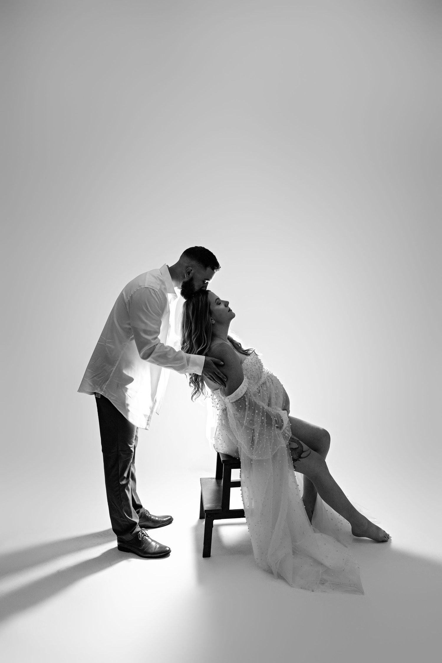 A romantic black and white studio portrait shows a couple in an intimate pose with dramatic lighting on a white backdrop.