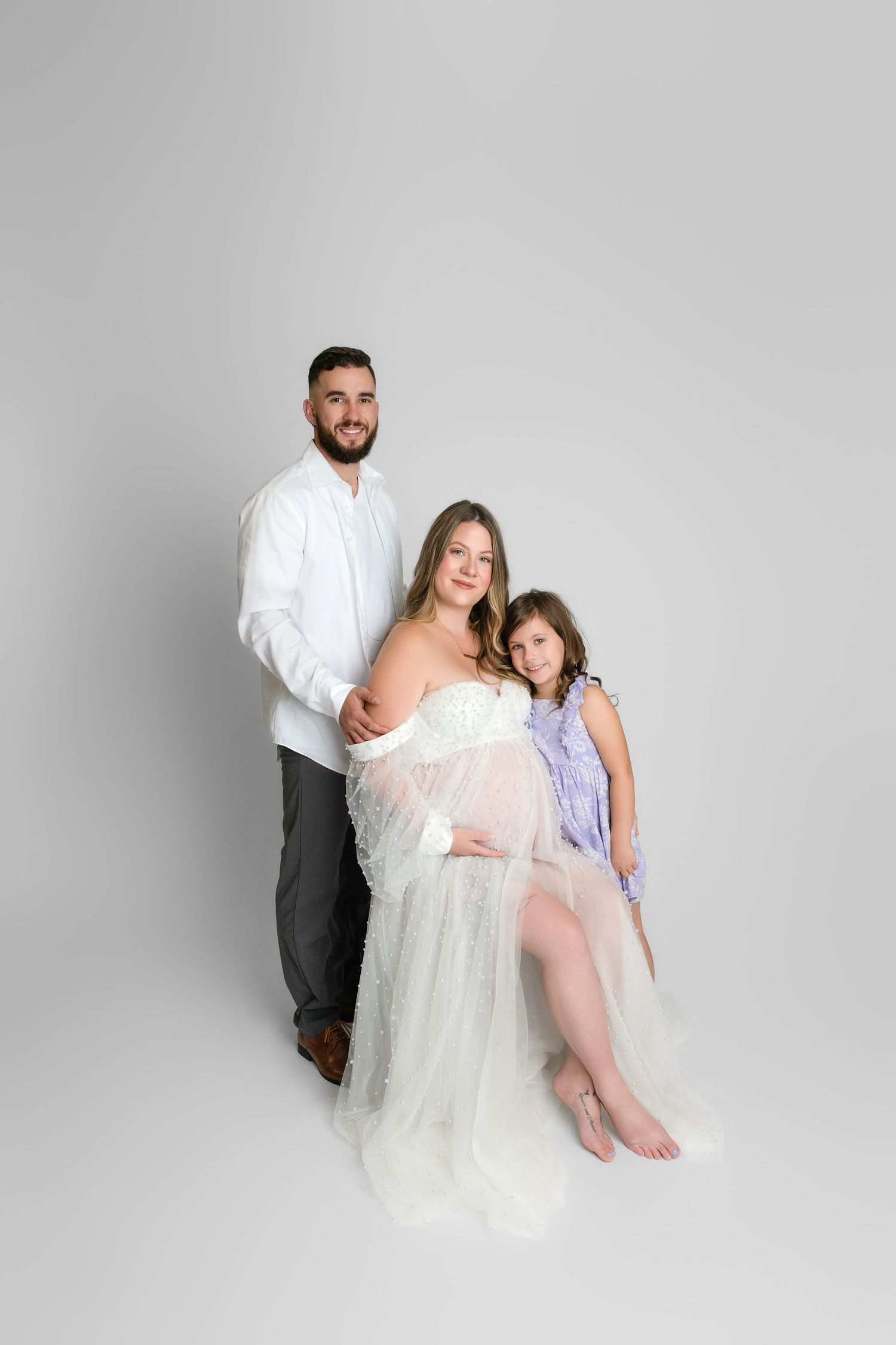 A family poses together for a maternity photo on a light gray backdrop, with the pregnant woman wearing a flowy white dress.