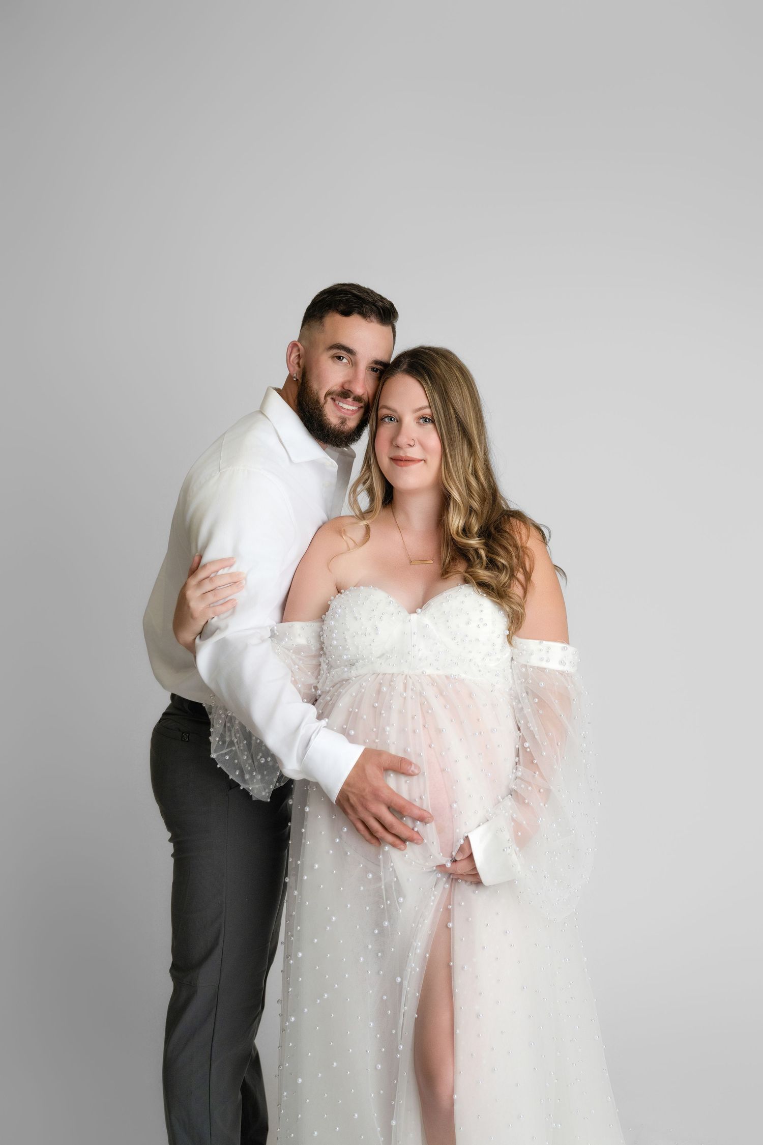 A romantic maternity photo shows a couple in elegant white attire embracing against a gray backdrop.