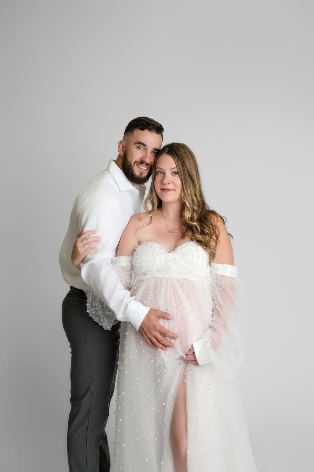 A romantic maternity photo shows a couple in elegant white attire embracing against a gray backdrop.