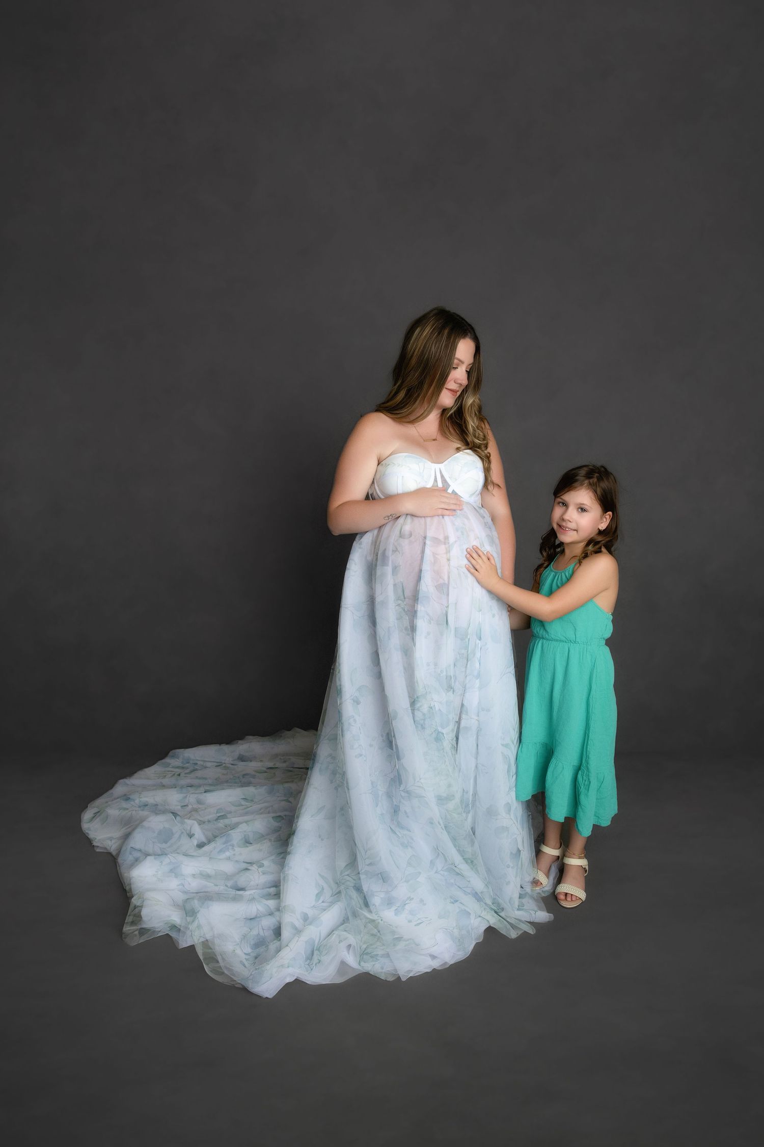 A pregnant woman in a flowing white gown stands next to a young child wearing a turquoise dress against a dark grey backdrop.