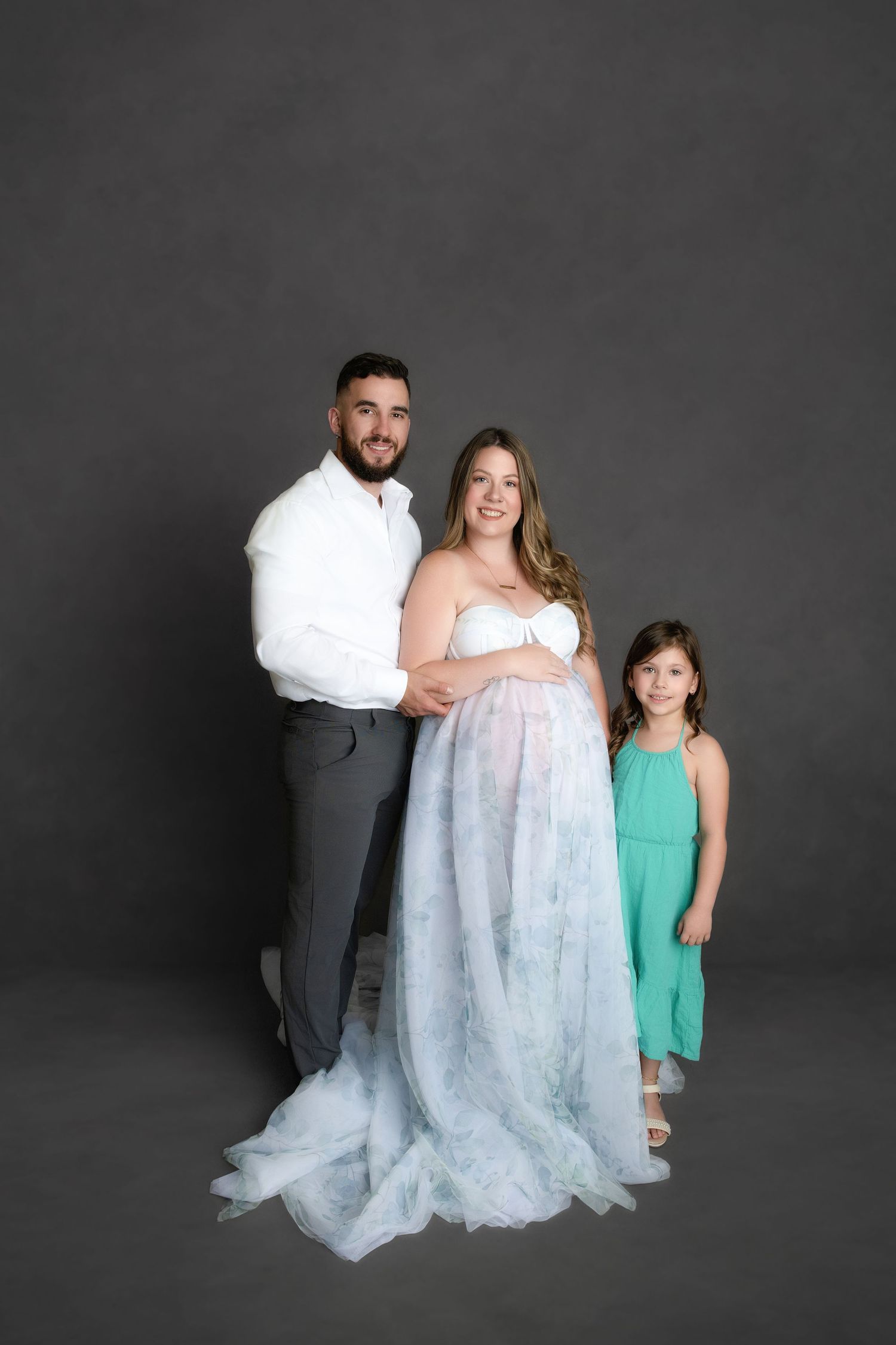 Studio portrait on dark gray background showing a family of three posing together in formal attire.