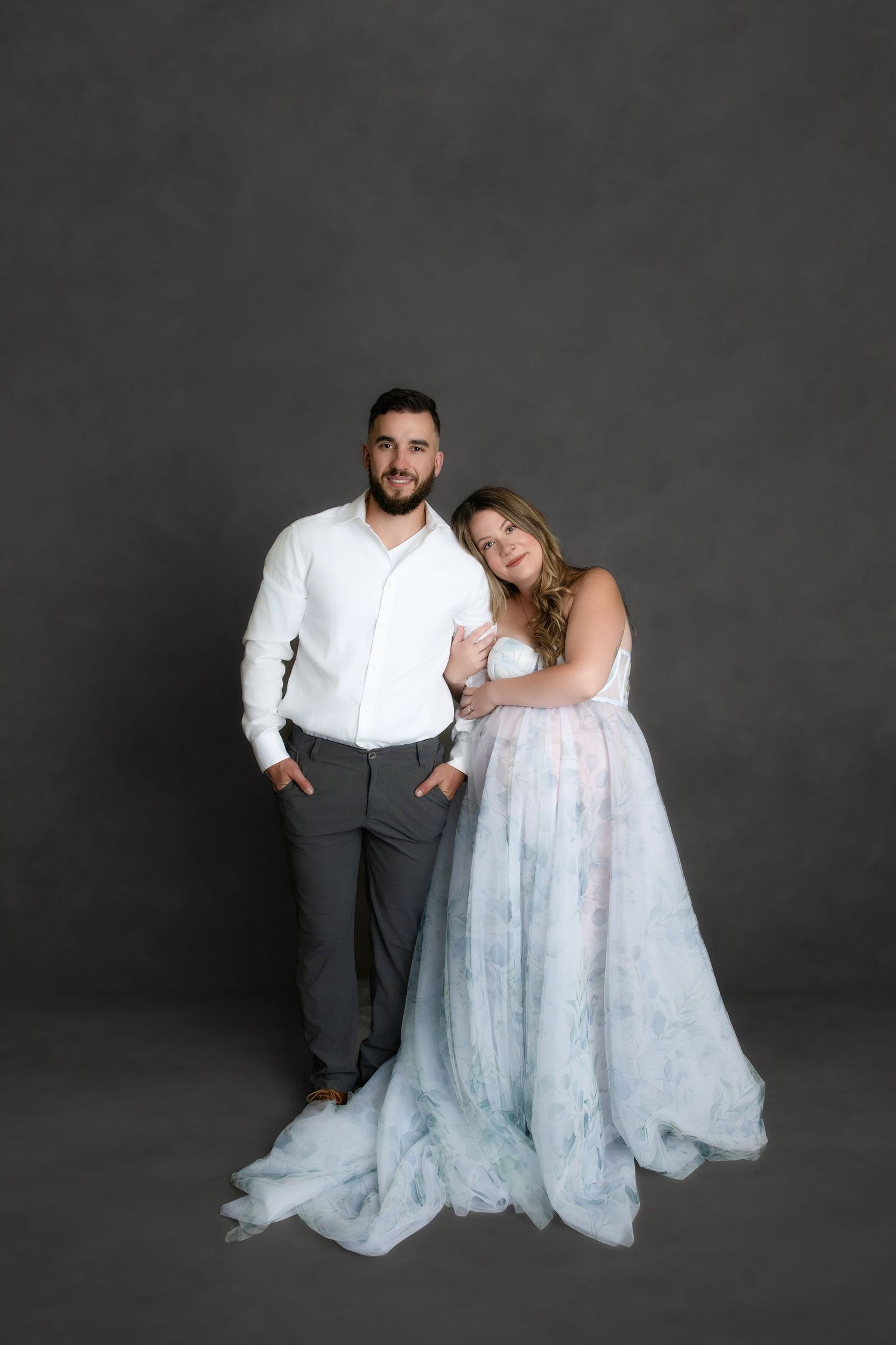 Romantic couple portrait session against dark gray backdrop with bride in flowing white lace gown and groom in dress shirt.
