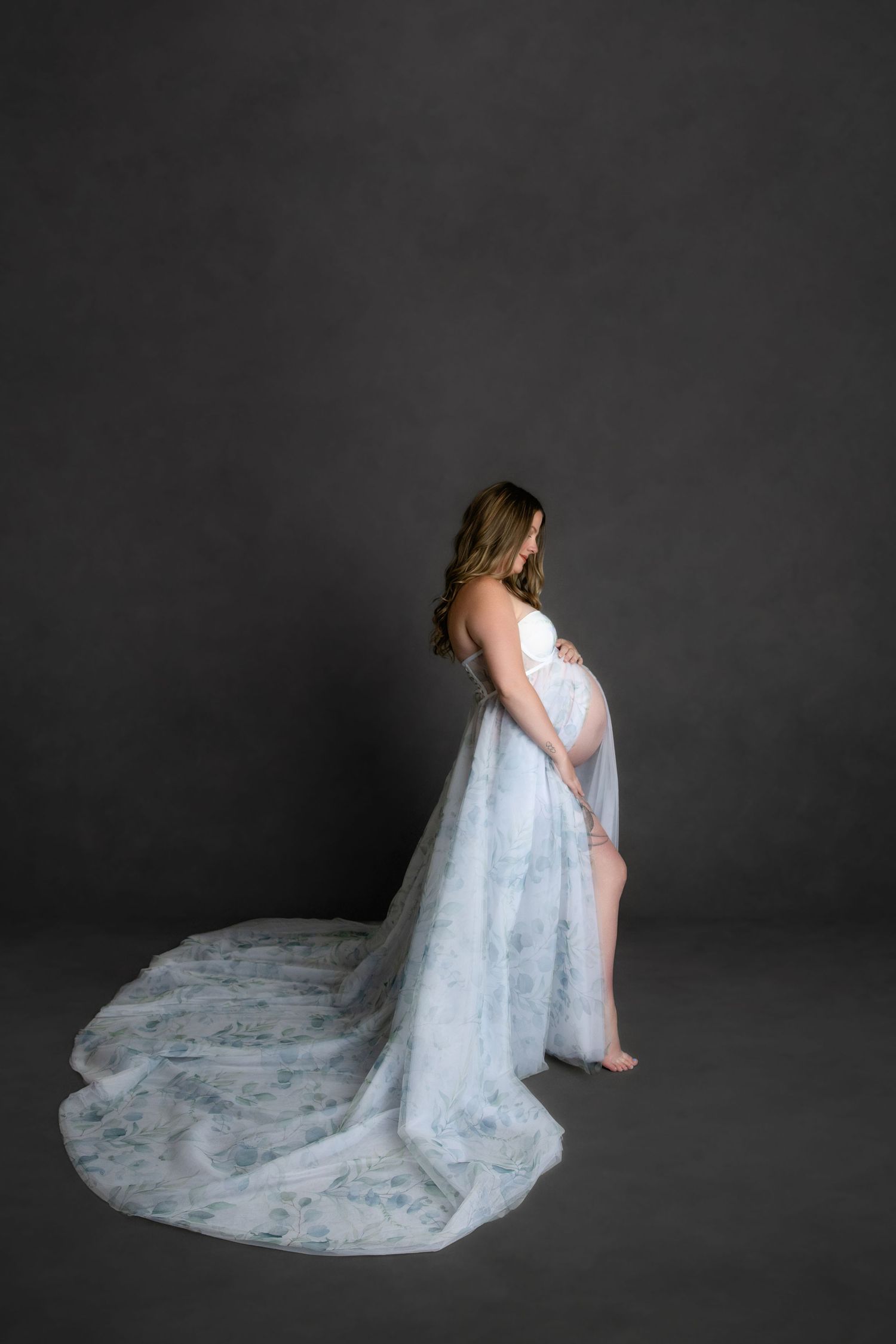 A pregnant figure in a flowing white lace gown stands in profile against a dark gray backdrop.