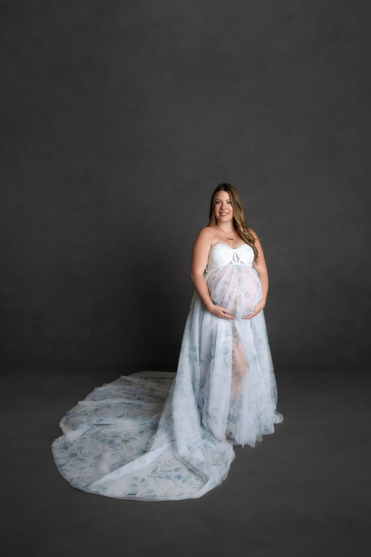 A pregnant woman in a flowing white lace maternity gown poses gracefully against a dark gray studio background.