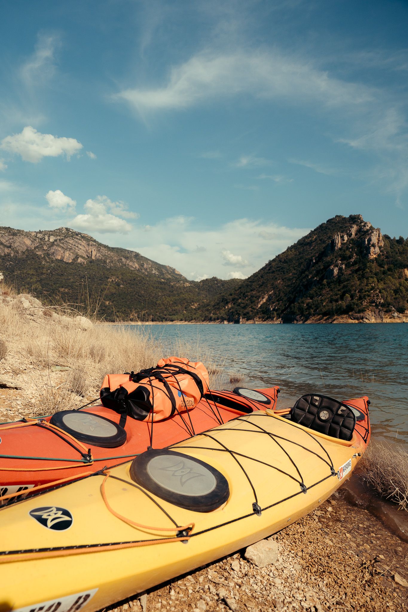 Orange and yellow kayaks rest on a rocky shoreline with mountains in the background.