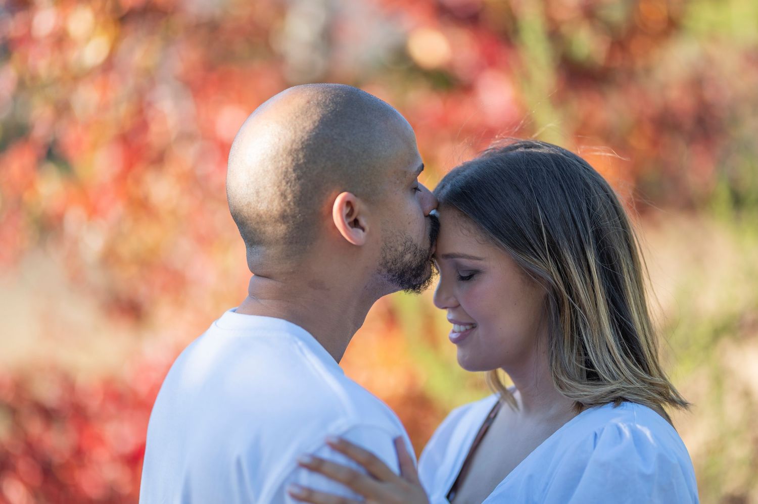 A romantic couple in white shirts embrace against a backdrop of vibrant autumn foliage with red leaves.