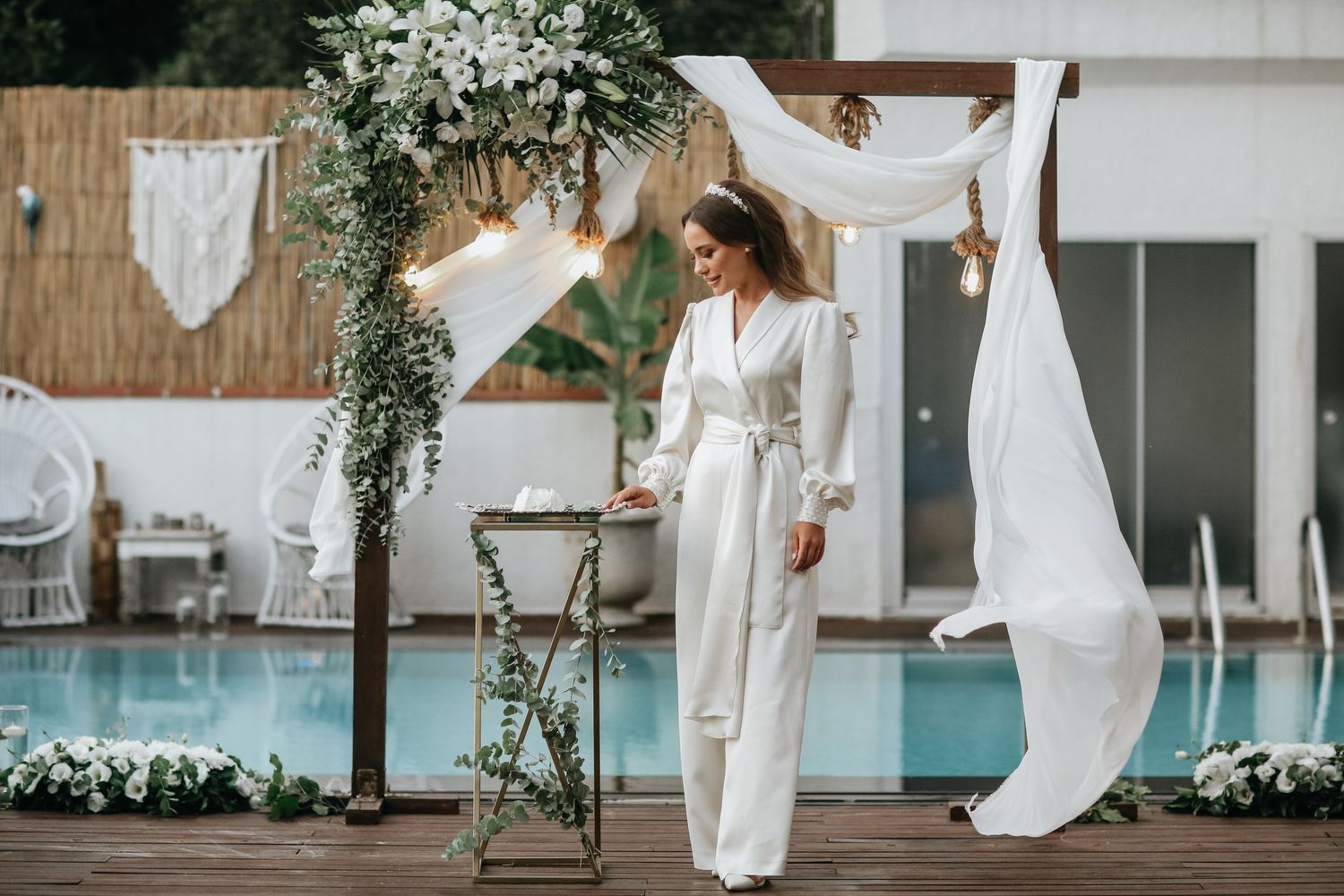 A modern minimalist wedding ceremony setup with white flowing fabric and greenery decorations next to poolside venue.