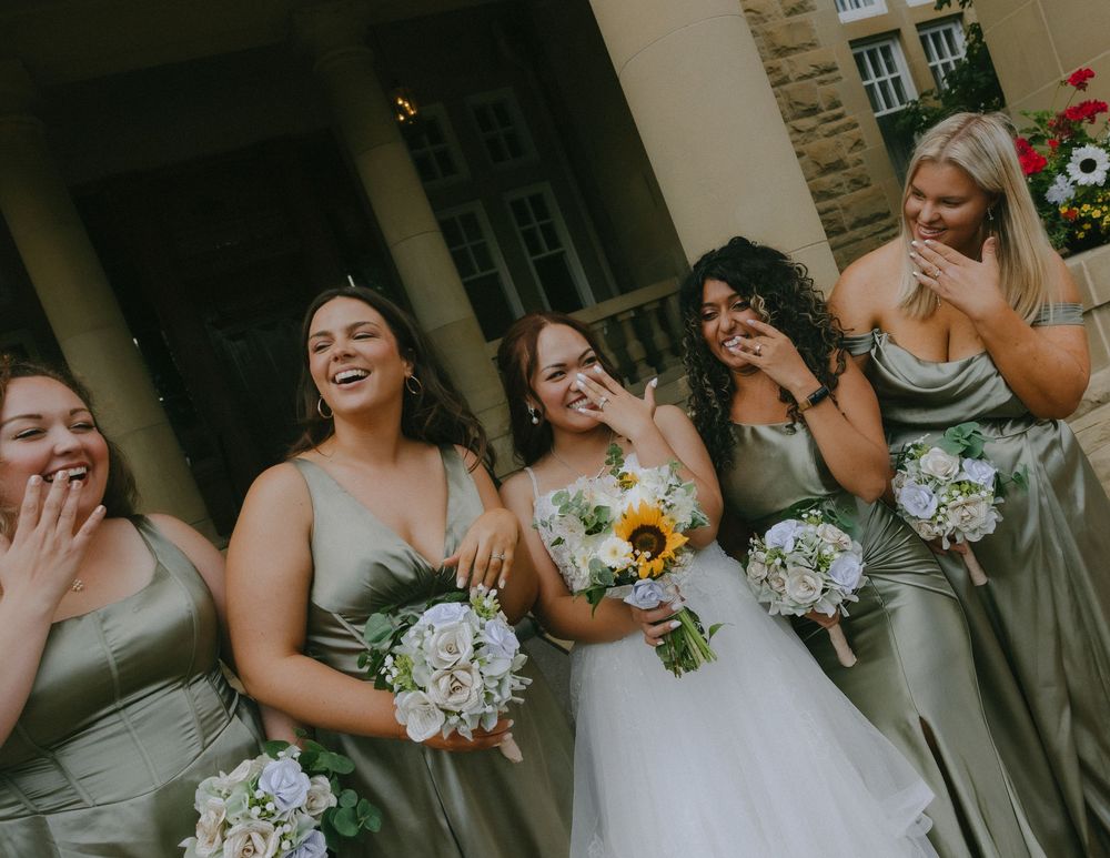 Wedding party laughs together while holding white and yellow floral bouquets in sage green bridesmaid dresses.