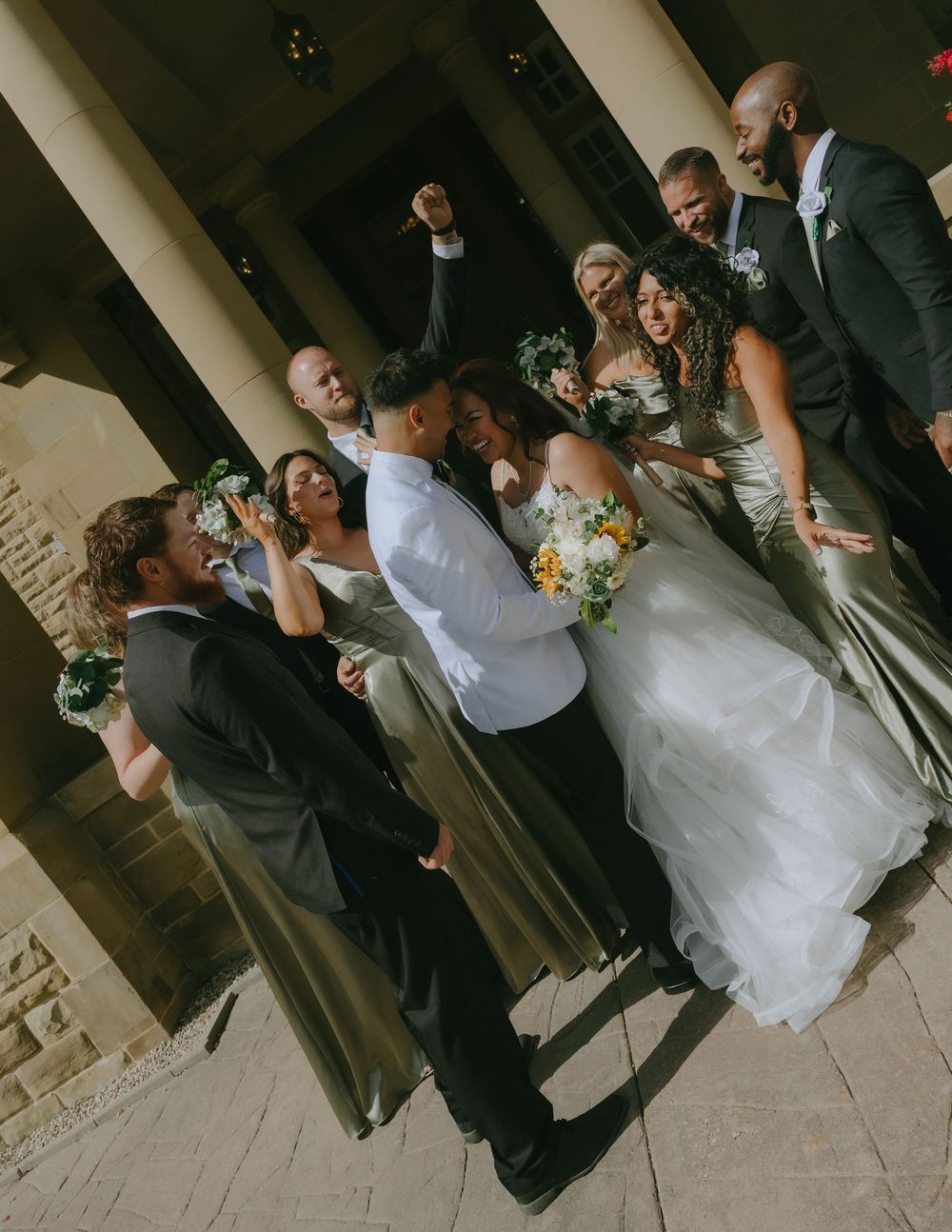 A joyful wedding party celebrates and poses together in a slanted architectural setting.