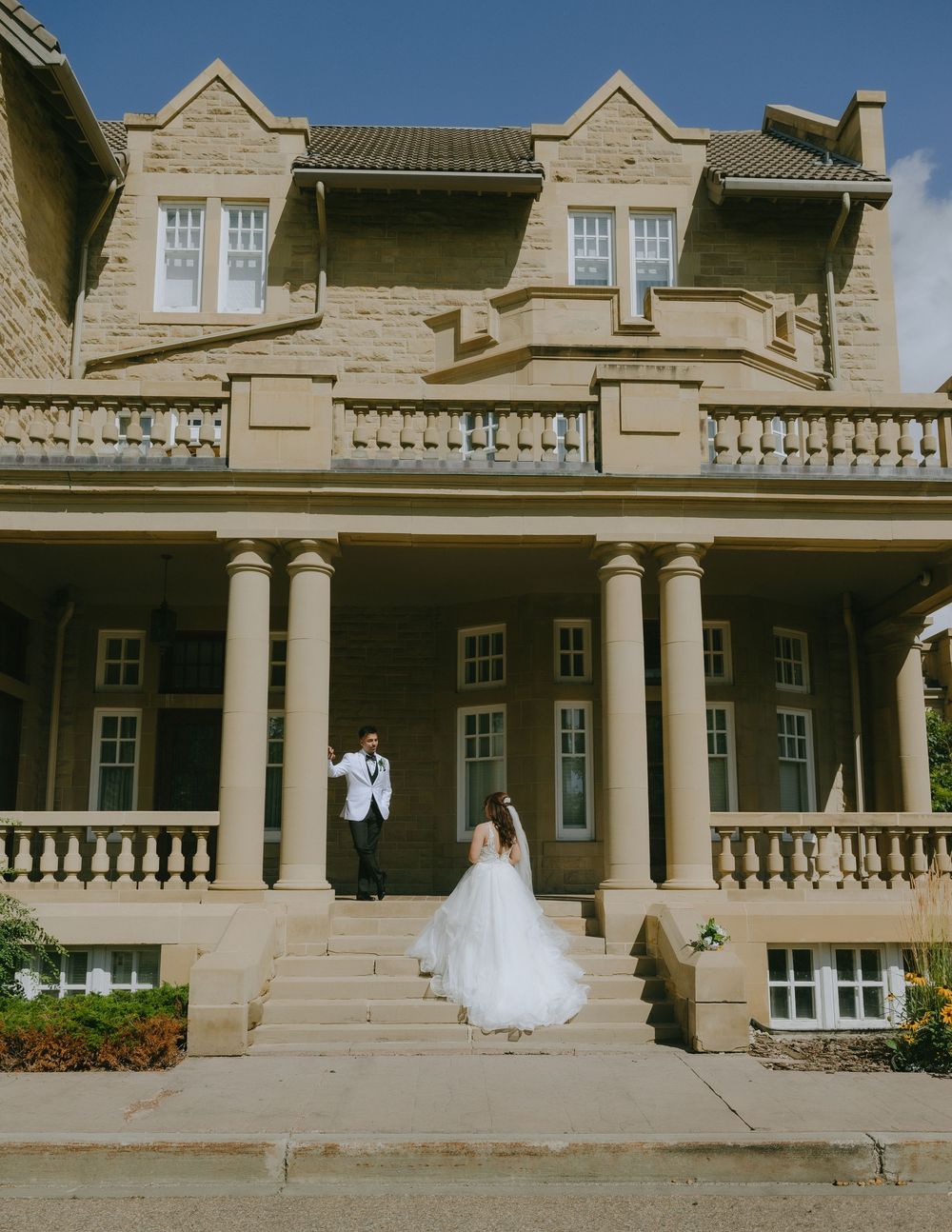 A wedding couple poses on the grand steps of a historic stone mansion with ornate columns and dormer windows.