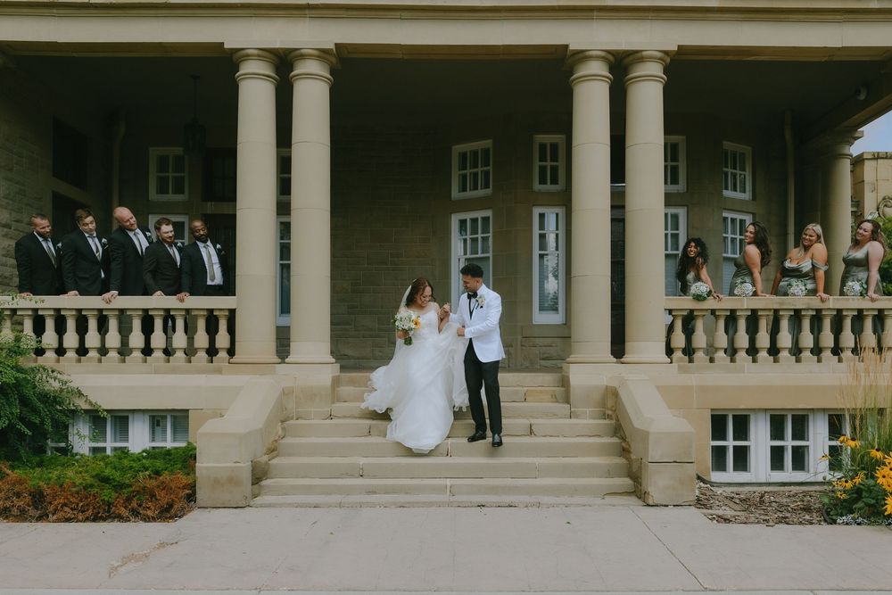 Wedding couple and guests pose on grand balcony of elegant colonial-style mansion during outdoor celebration.