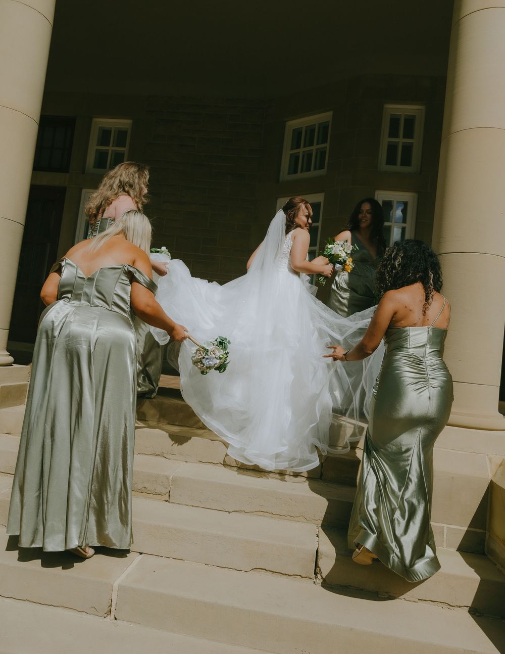 Bridesmaids in silver satin dresses help carry a bride's flowing white wedding gown up stone steps.