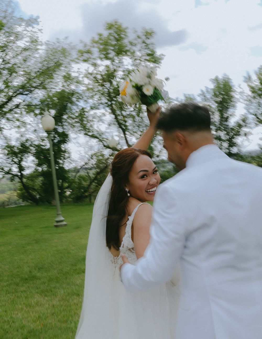 A joyful couple celebrating their wedding day outdoors in a green garden setting under cloudy skies.