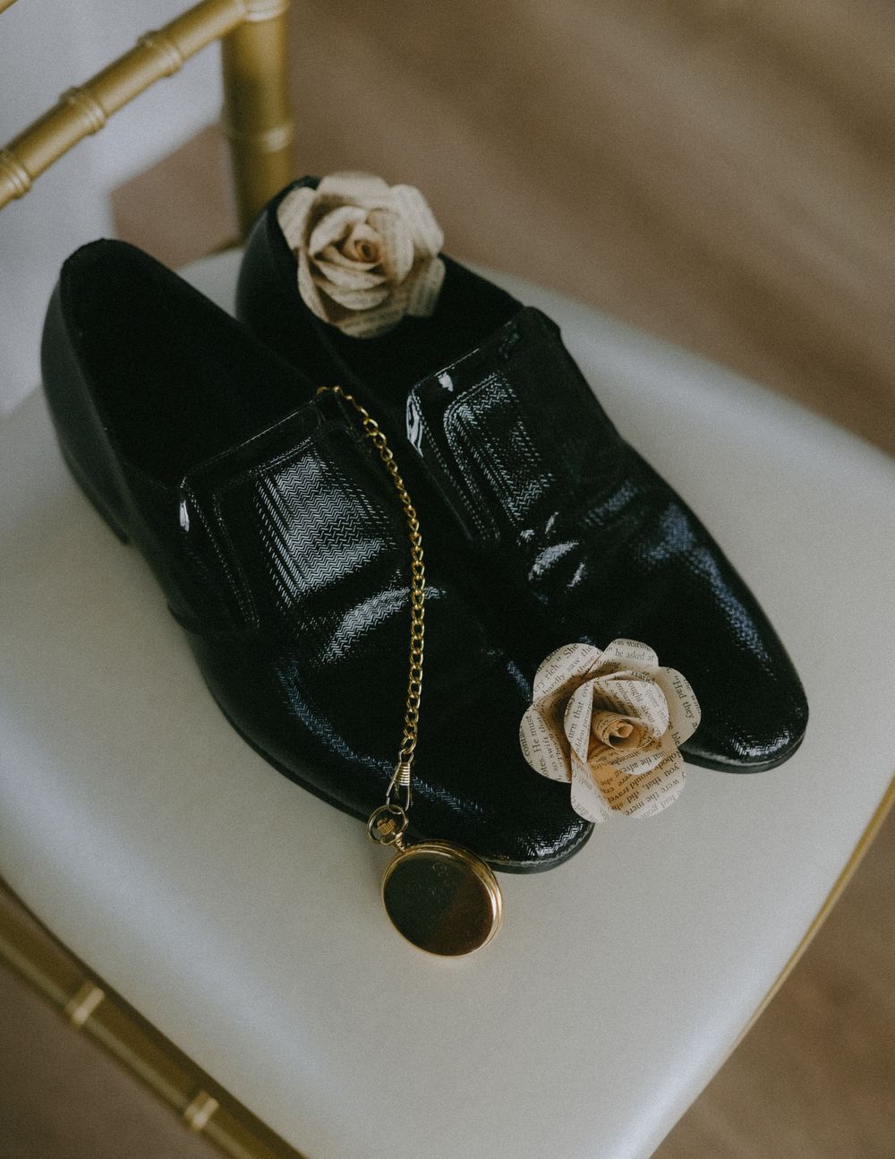 Black leather loafers with a delicate white rose and gold chain displayed on white surface in soft lighting.