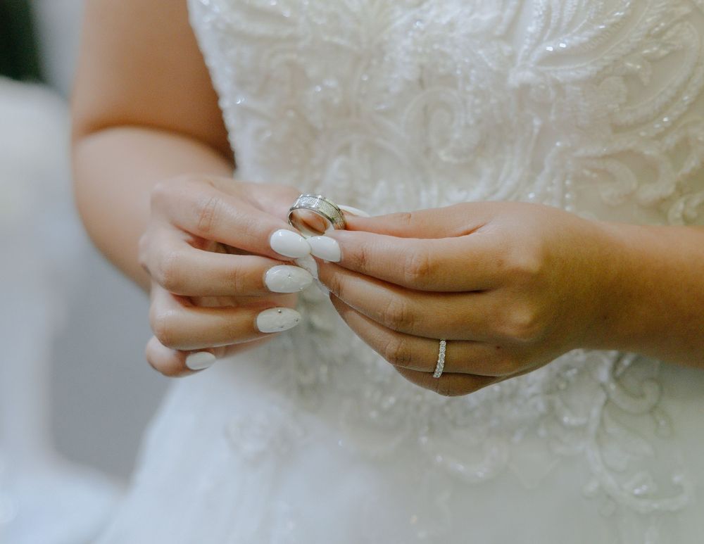Hands with white manicured nails hold a wedding ring against a lacy white dress background.