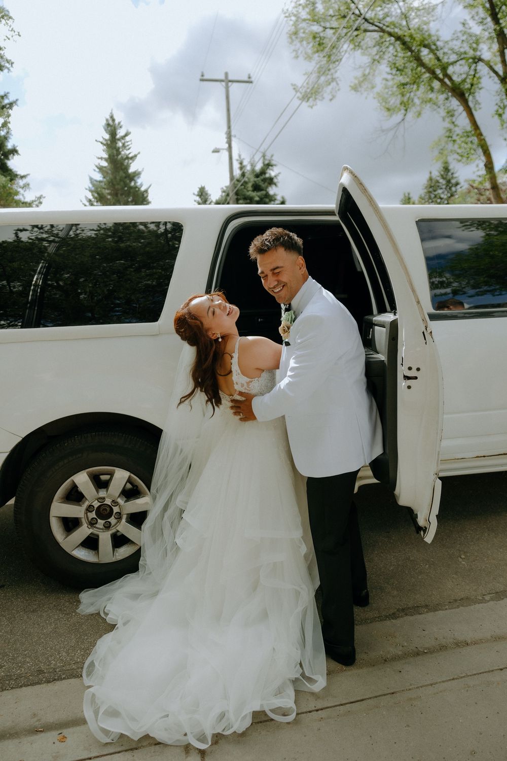 Bride and groom share a playful moment by their white limousine after their wedding ceremony.