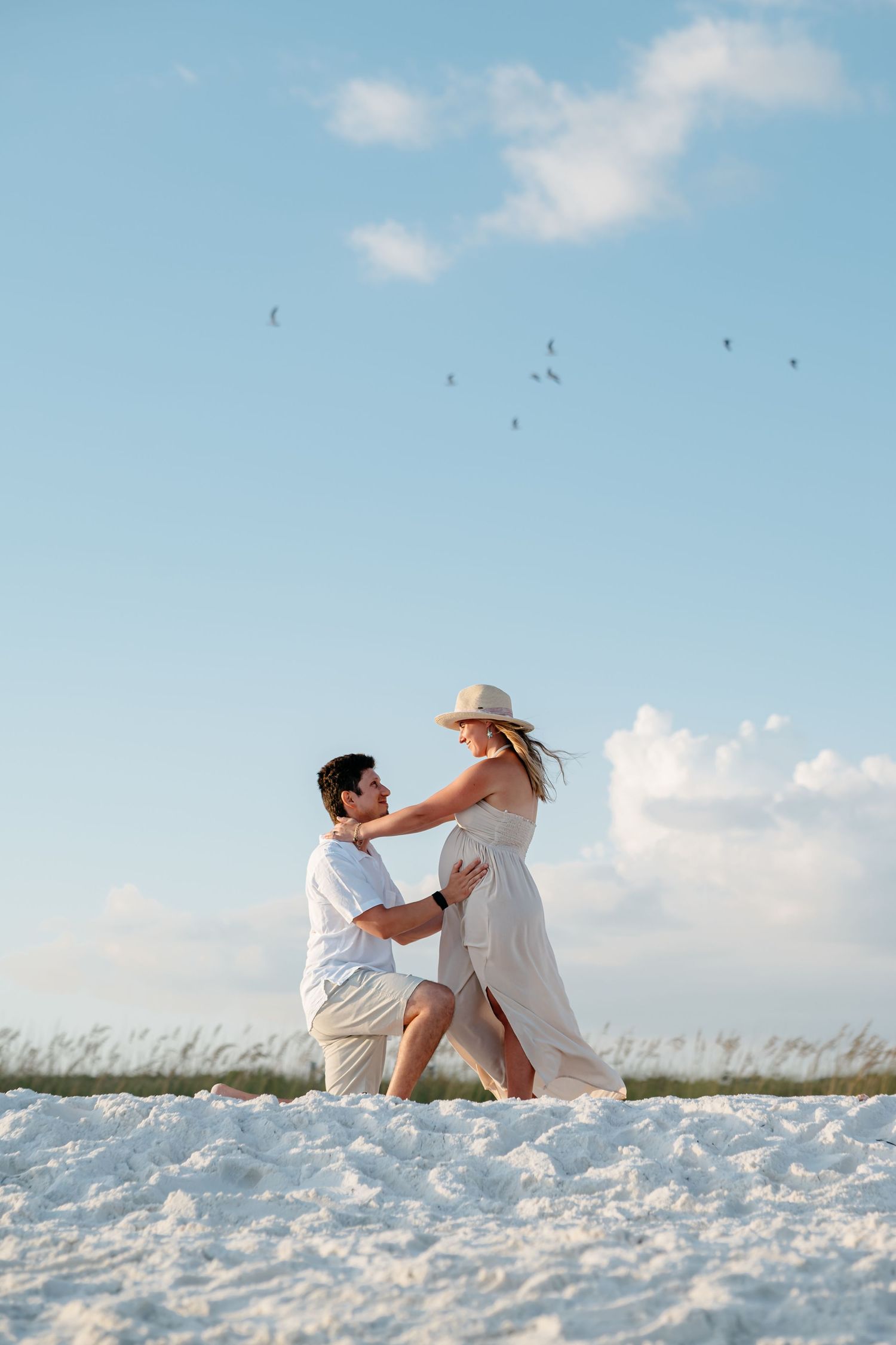 A romantic beach proposal moment captured on white sand with birds flying in the blue sky.