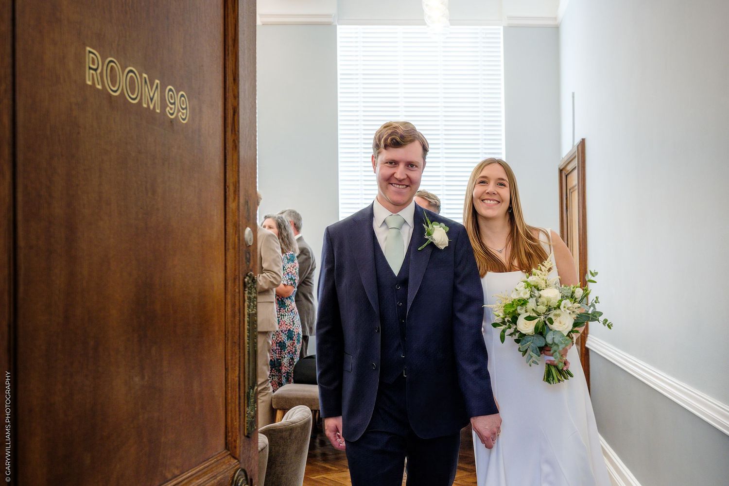 A joyful couple stands together in a hallway as the bride holds a white floral bouquet.