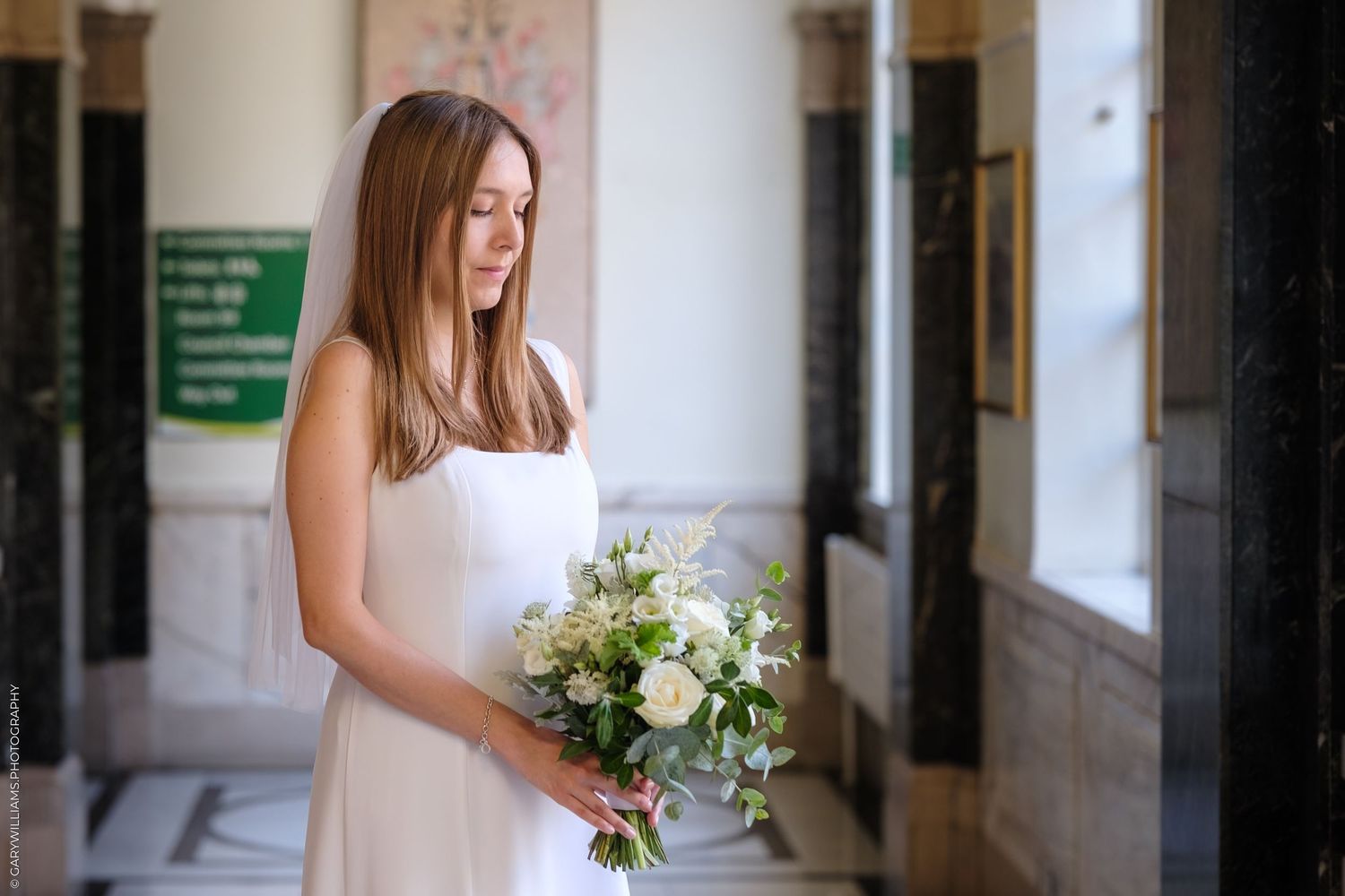 A bride in a white wedding gown holds a bouquet while walking down a bright corridor with white walls.