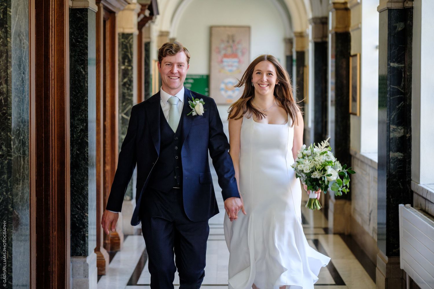 A bride and groom walk through an elegant arched hallway with traditional architectural details and wooden doors.