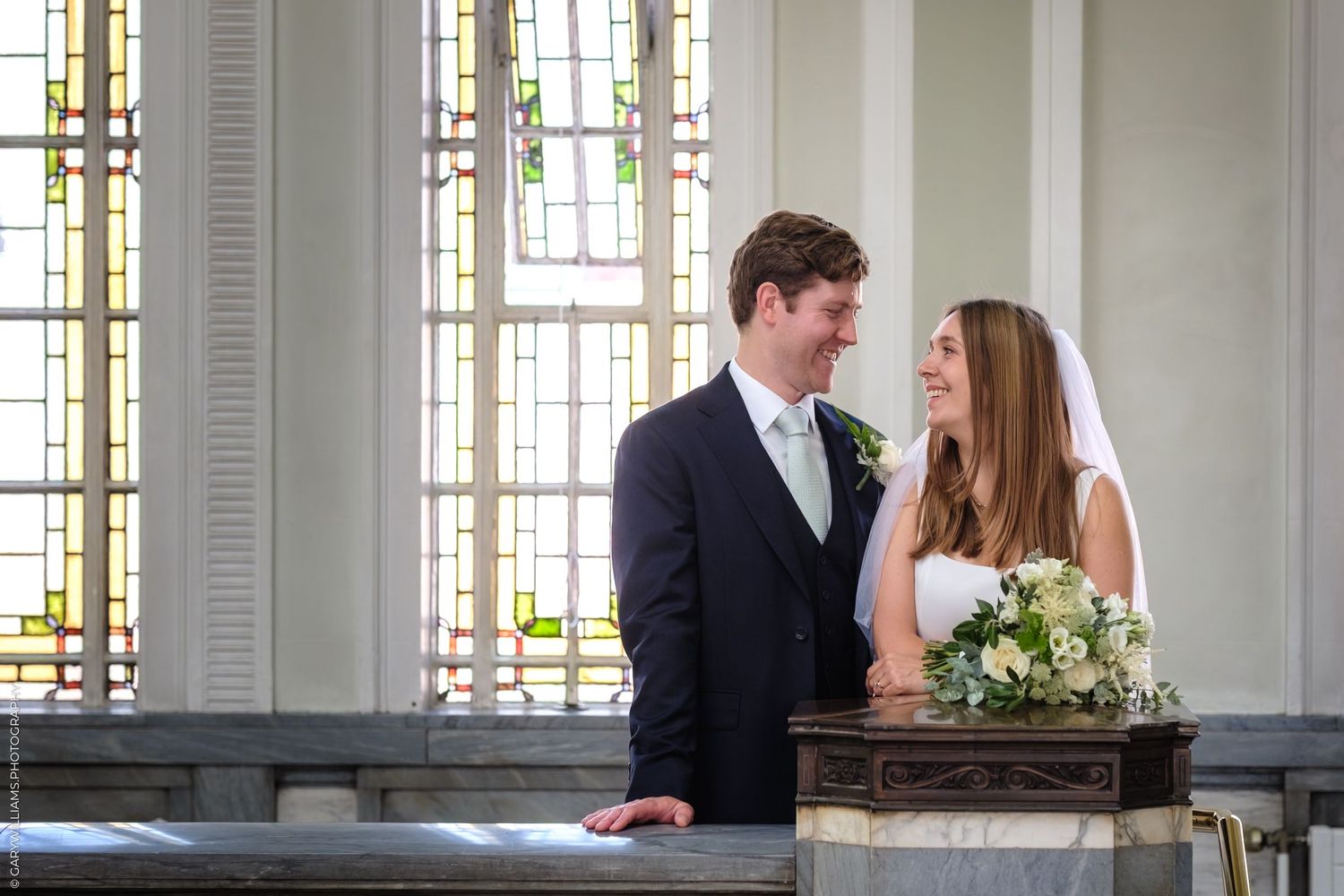 A couple kisses during their wedding ceremony in an elegant white room with tall windows and decorative columns.