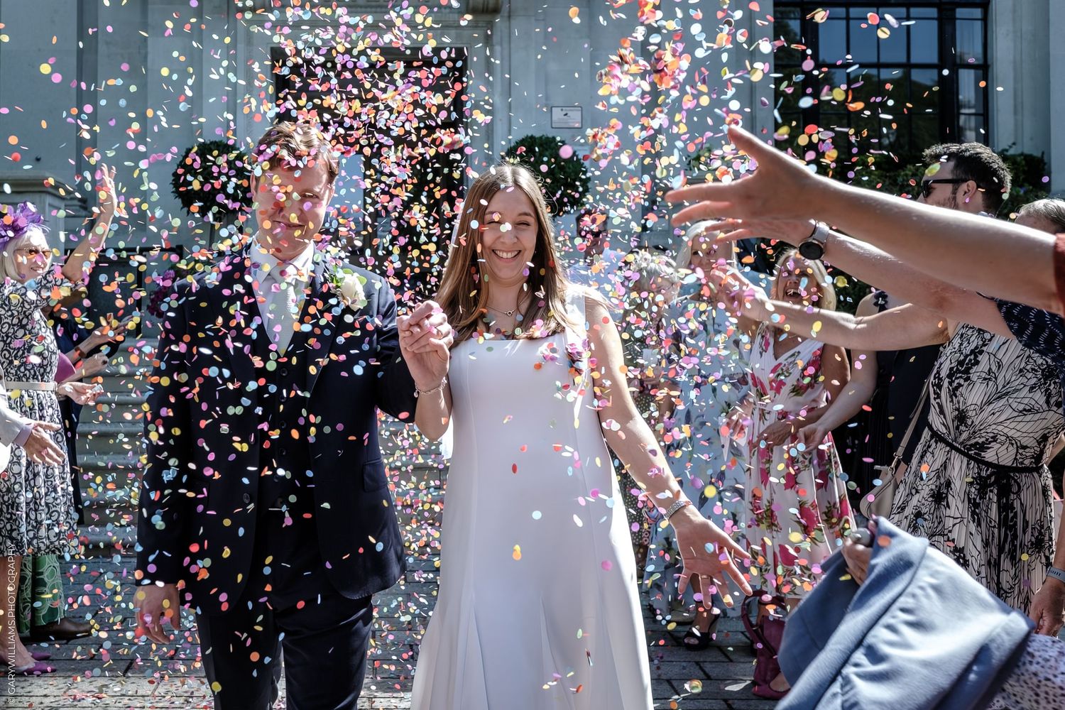 Wedding couple walks through joyful burst of confetti outside a building after ceremony celebration.