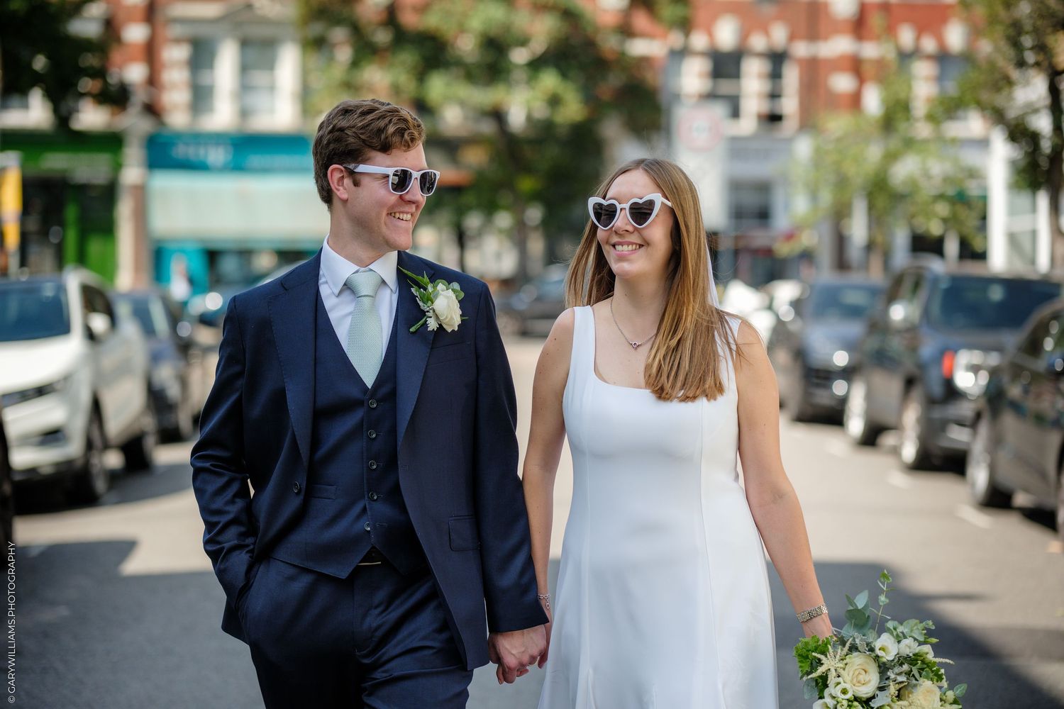 A happy couple walking down a street on their wedding day wearing a navy suit and white dress with sunglasses.