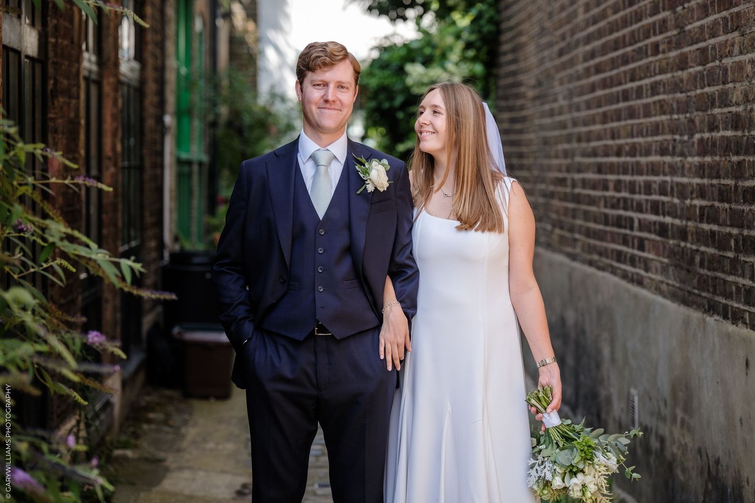 A couple stands in an urban alleyway wearing formal attire with a brick wall backdrop.