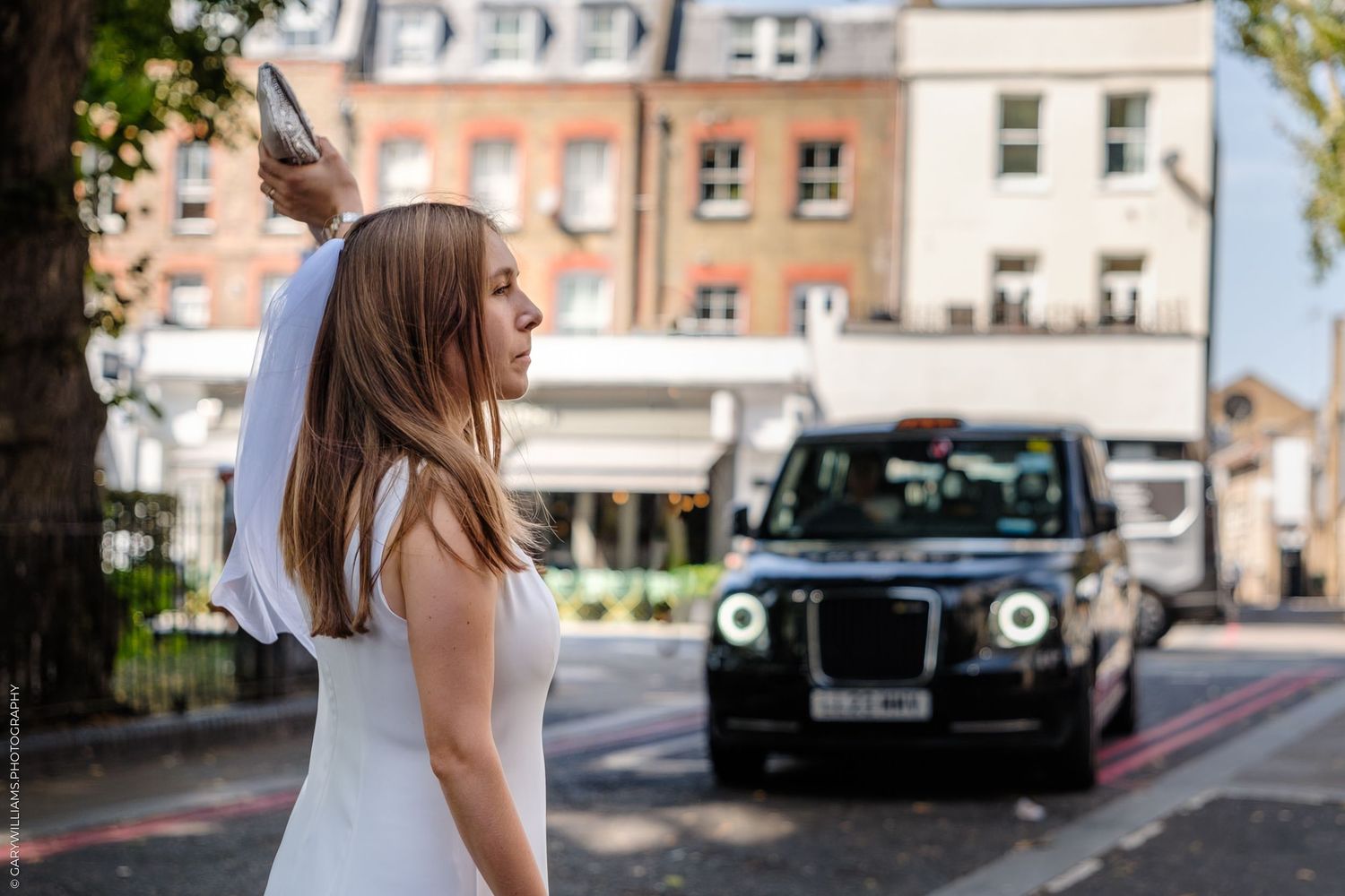 A bride standing in a white wedding dress near parked vehicles on a city street during the daytime.